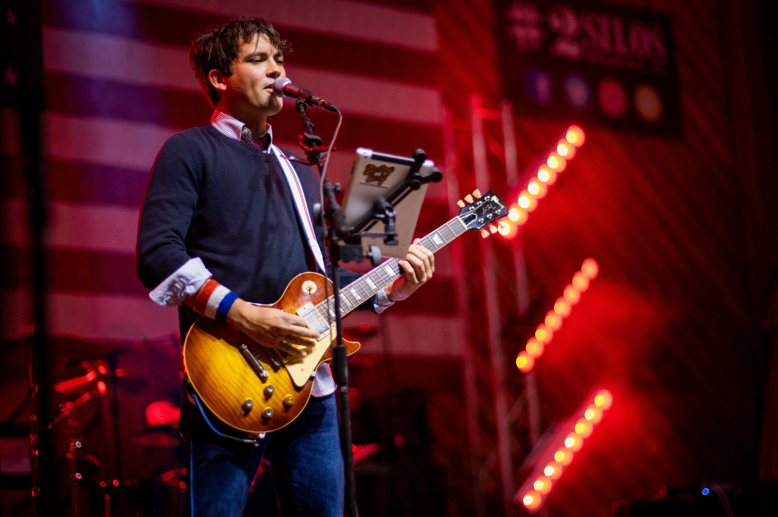 Young man playing guitar and singing on stage with red lighting and striped background.