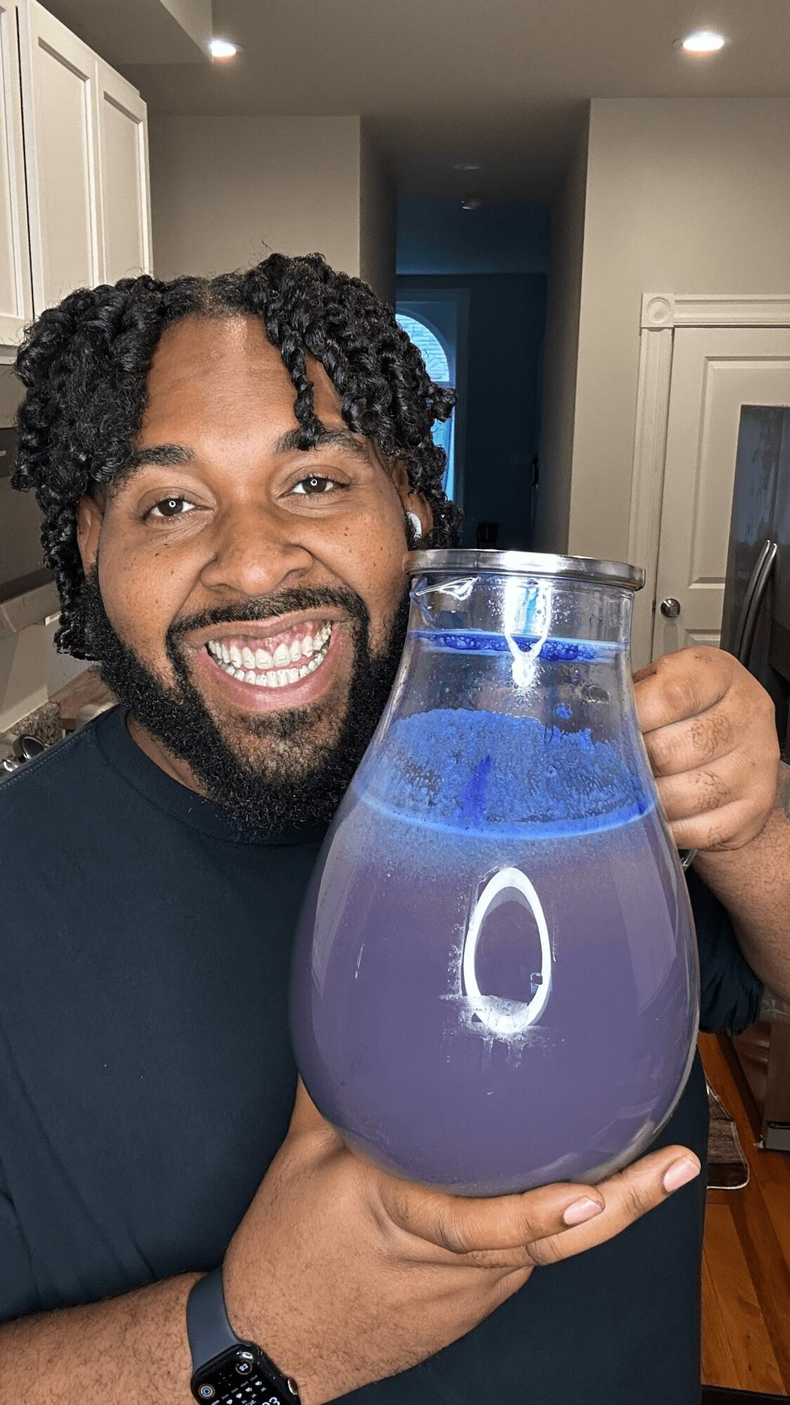 Person smiling holding a large glass pitcher with purple liquid, in a kitchen with white cabinets and a doorway in the background.