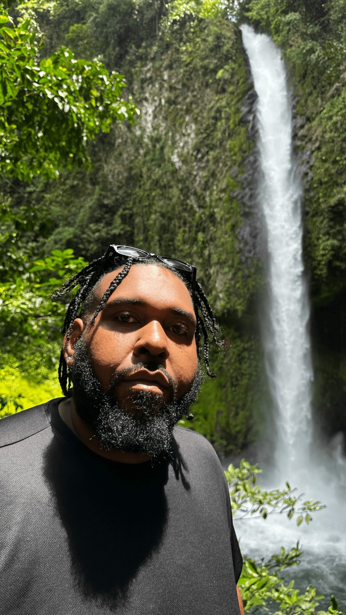 Man with braided hair and beard in front of a waterfall surrounded by greenery.