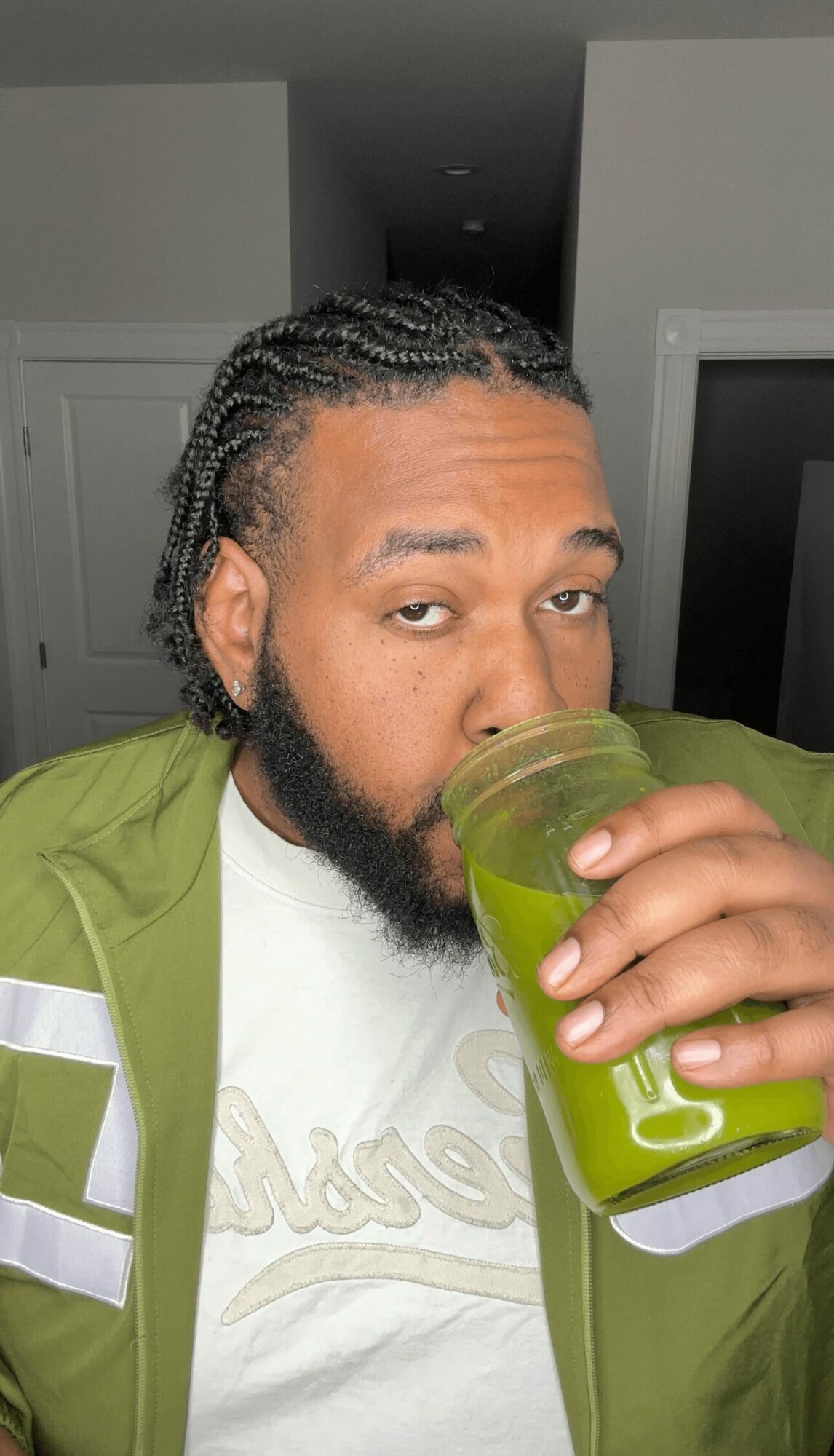 Person with braided hair and beard holding a green drink in a clear cup, wearing a green jacket and white shirt.