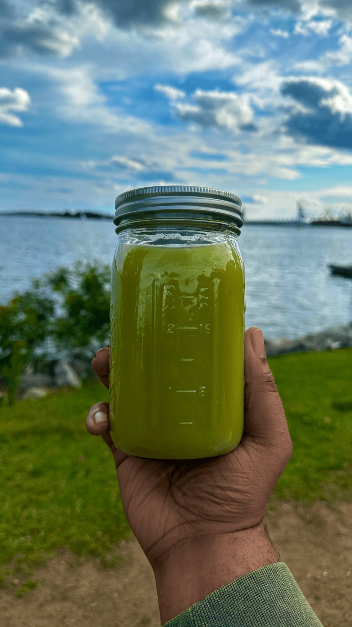 Hand holding a jar of green juice against a lakeside background with blue sky and clouds.