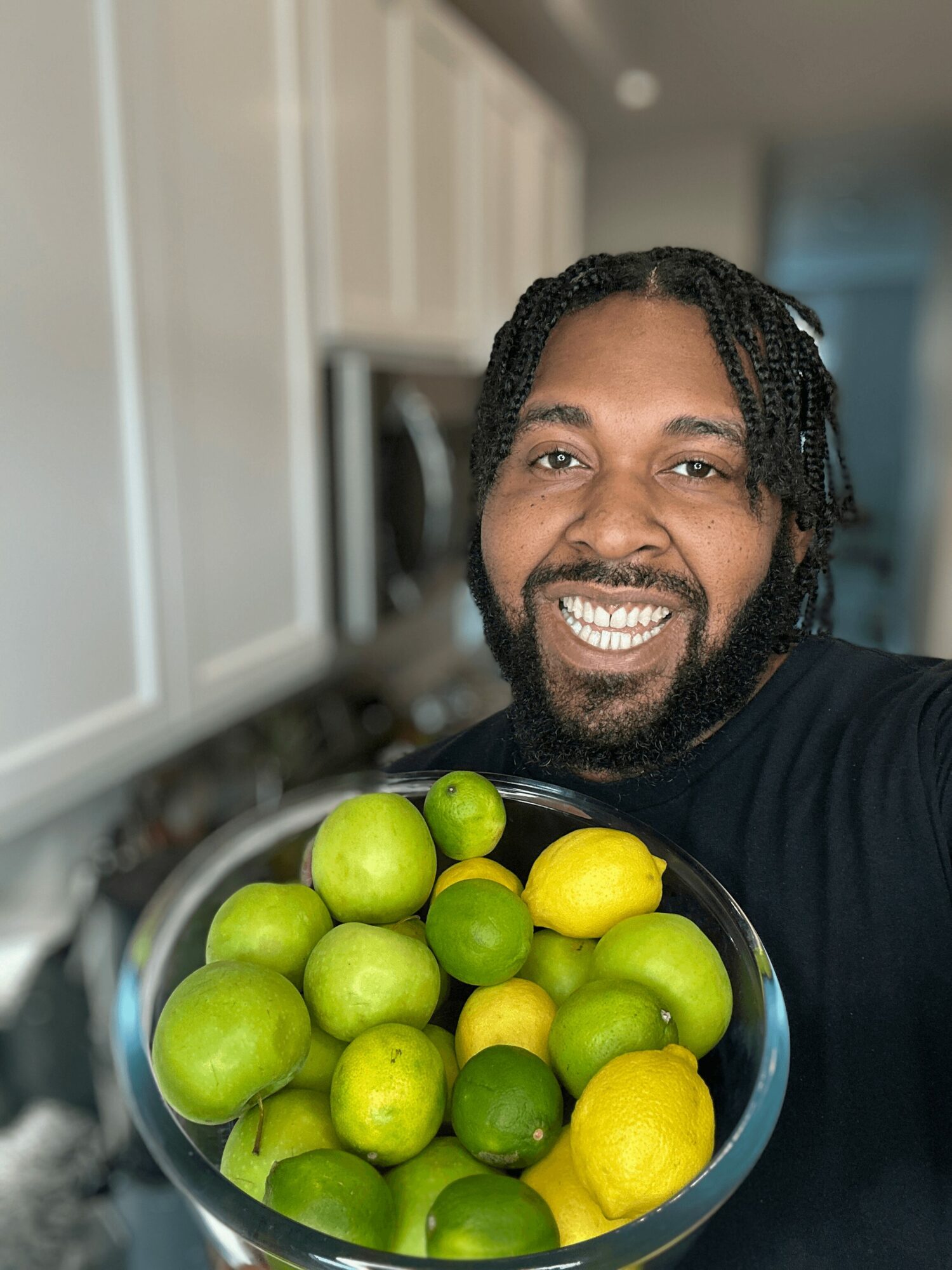 Person with dreadlocks smiling, holding a bowl of green and yellow lemons in a kitchen.
