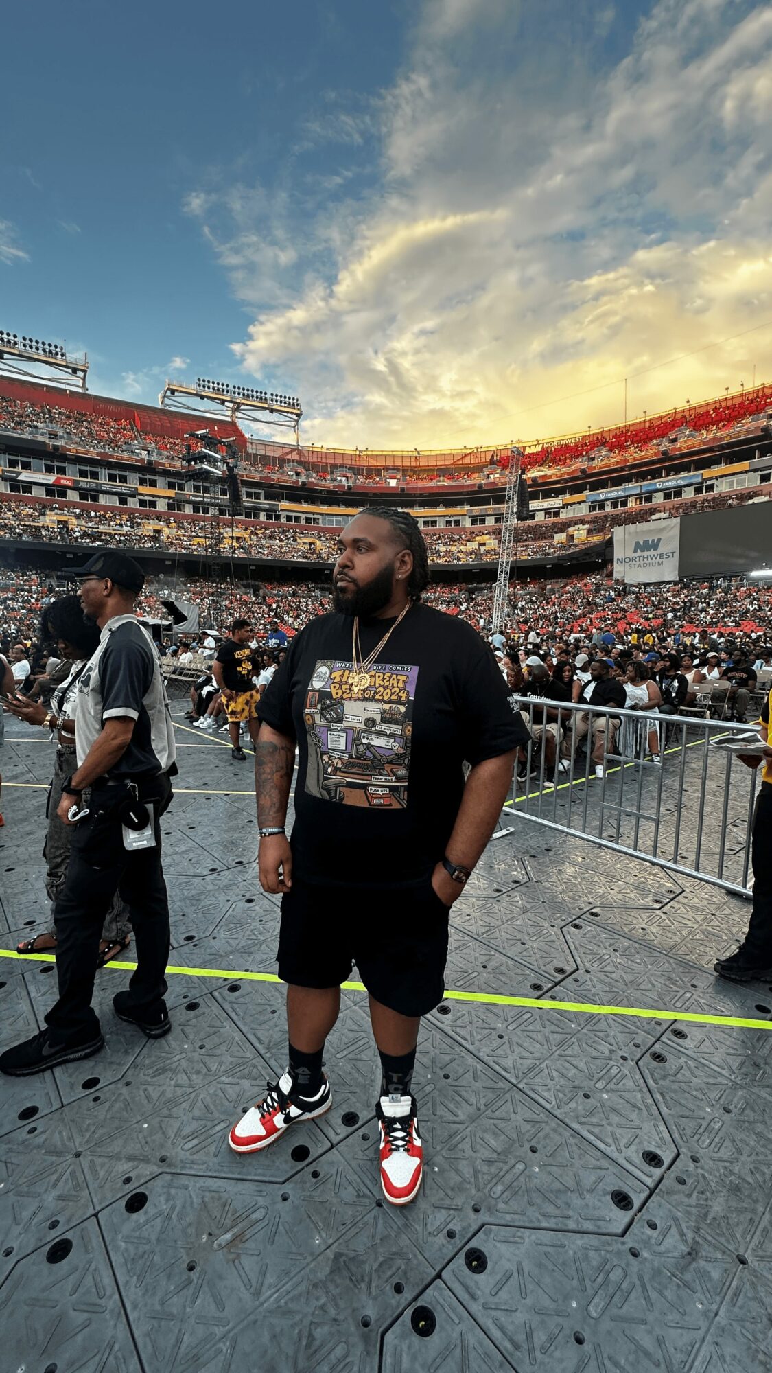 Person standing in a stadium with a large crowd and sky in the background, wearing a black t-shirt and shorts.
