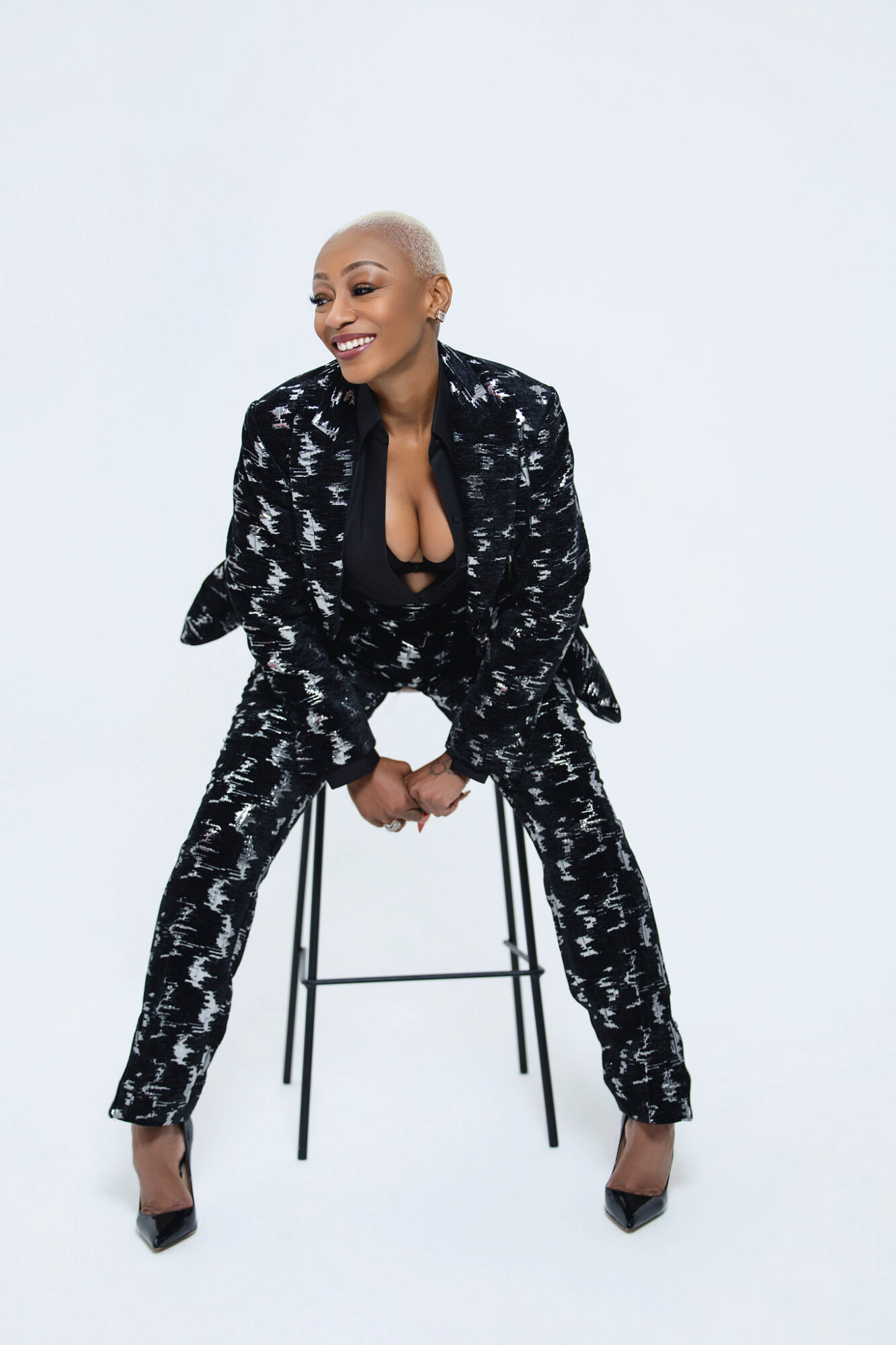 Woman with short blonde hair sitting on a stool, smiling, wearing a black and white patterned suit with high heels.