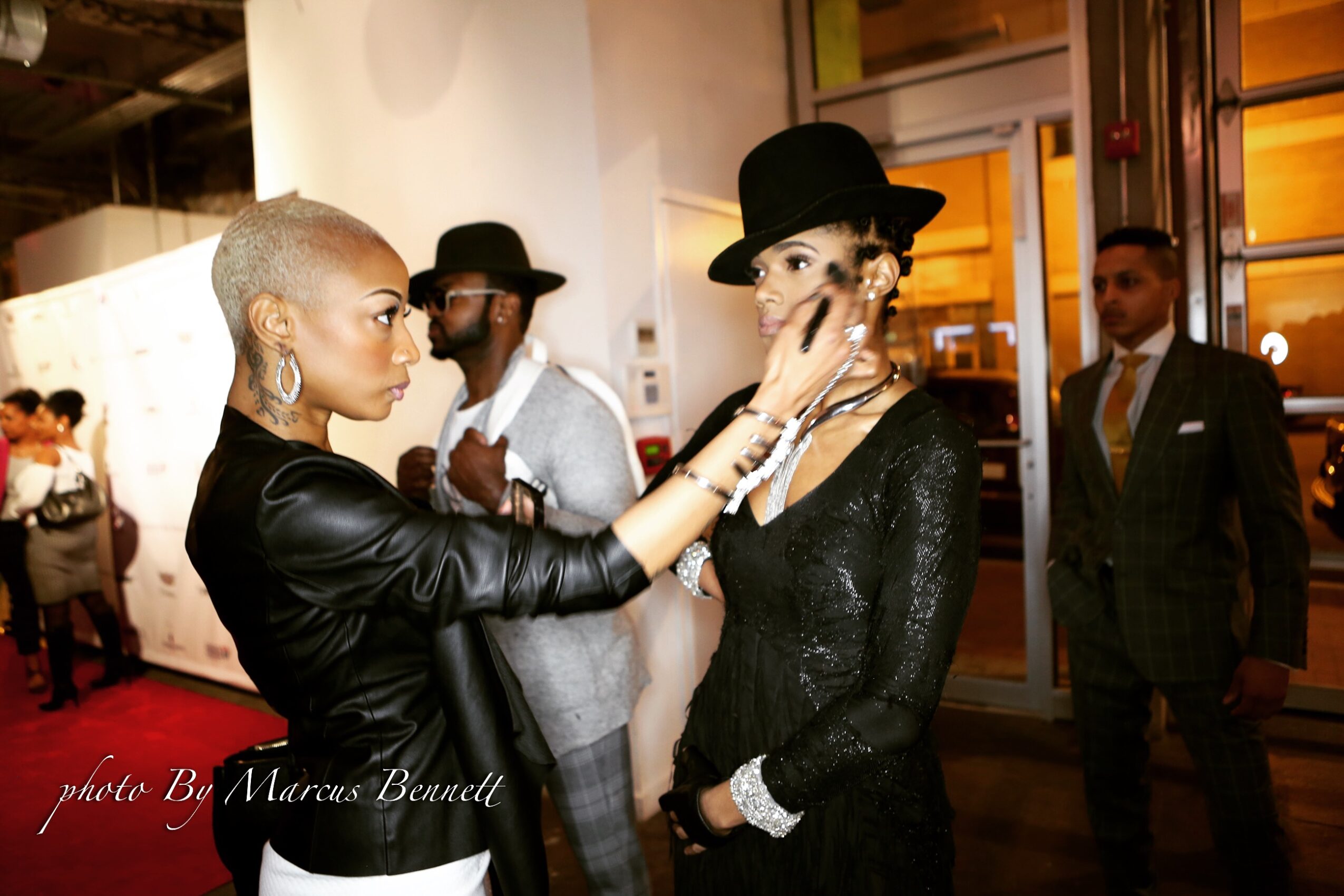 Two women with short hair and earrings face each other, one touching the other's face, in an indoor event setting.