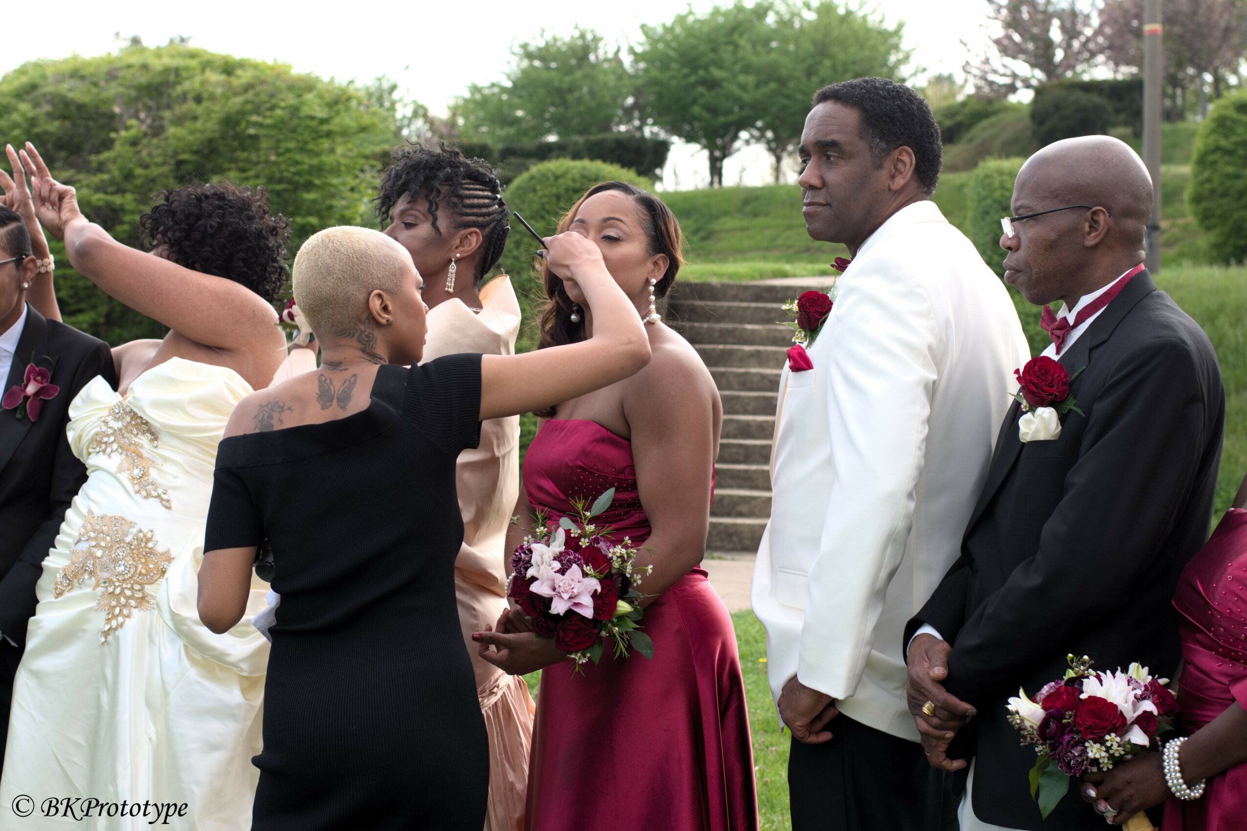 Group of people at a wedding ceremony outdoors, with a woman in a red dress and others in formal attire, some with flowers.