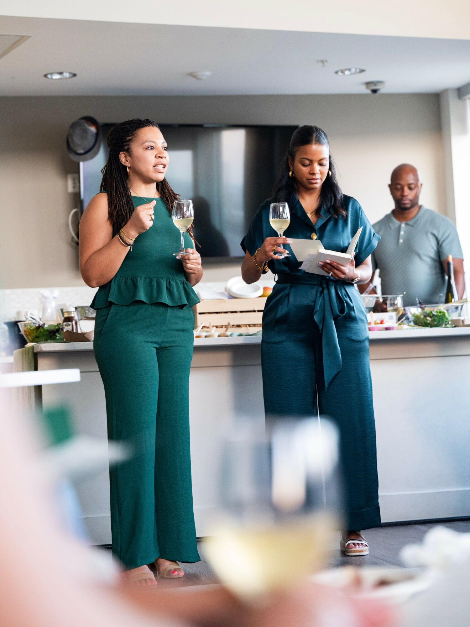 Two women stand in a kitchen, holding wine glasses, one reading from a tablet, with a man in the background.