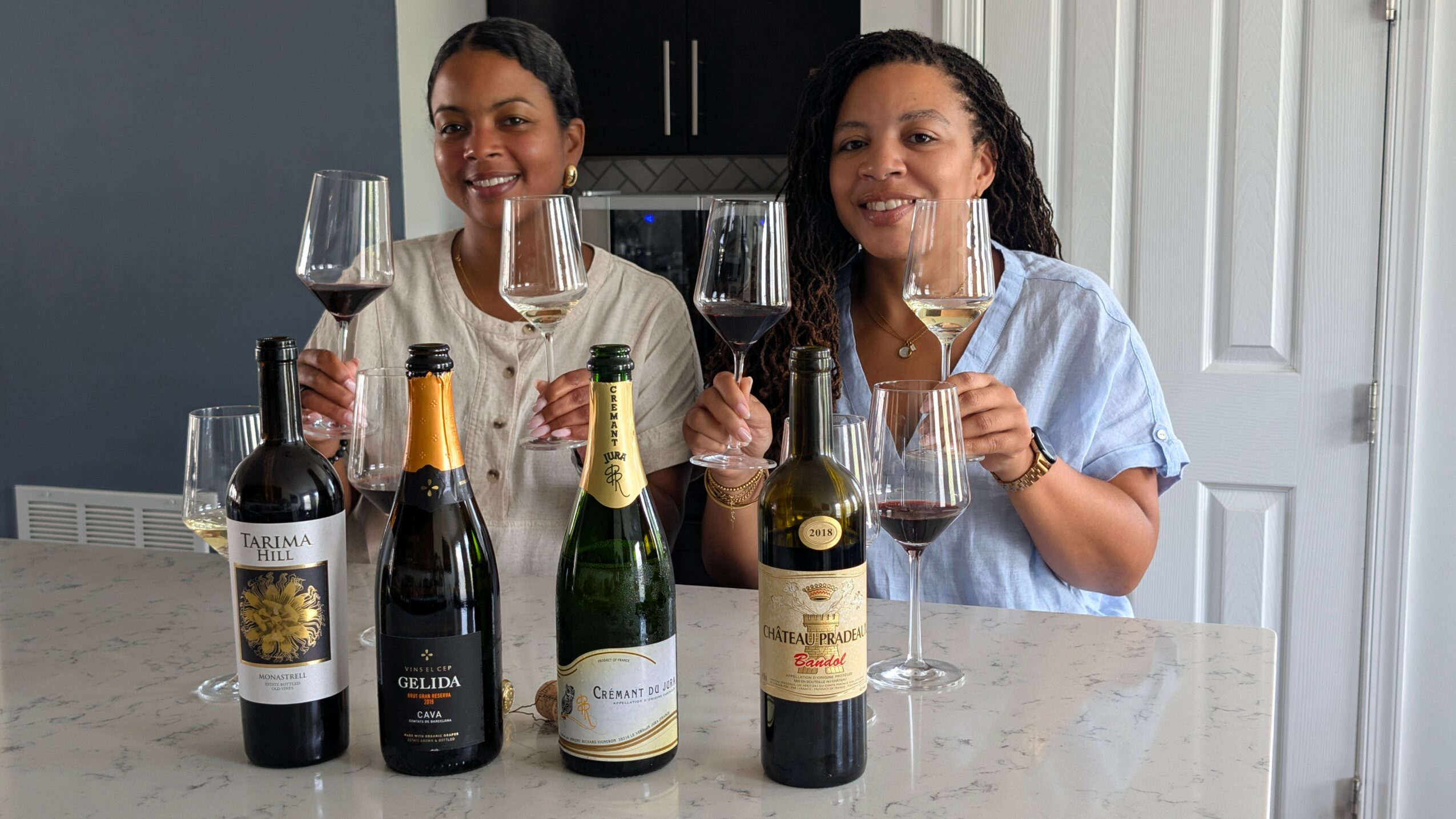 Two women smiling and holding wine glasses, standing behind a counter with four bottles of wine.
