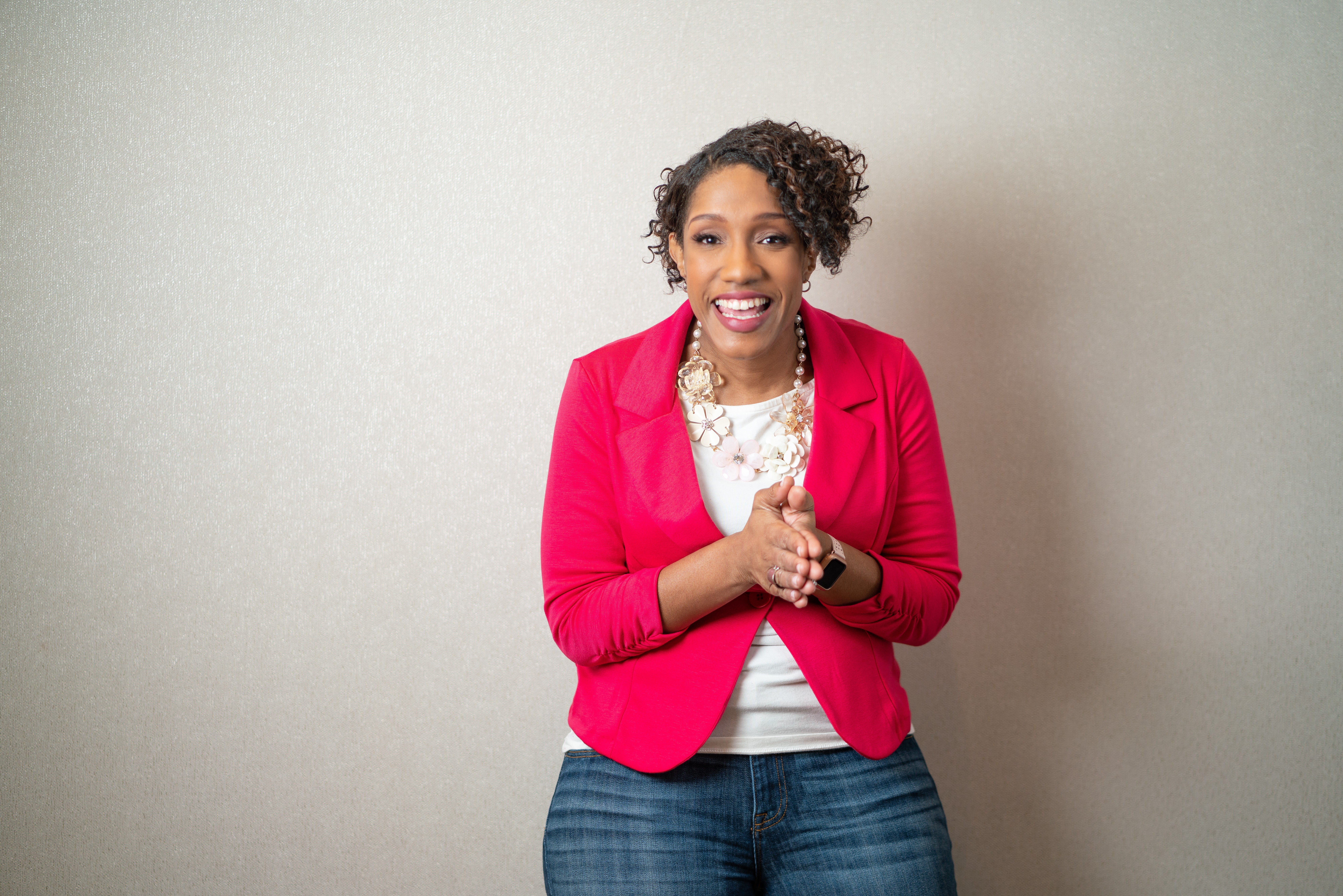 Smiling woman with curly hair wearing a red blazer and jeans, standing against a plain light background.