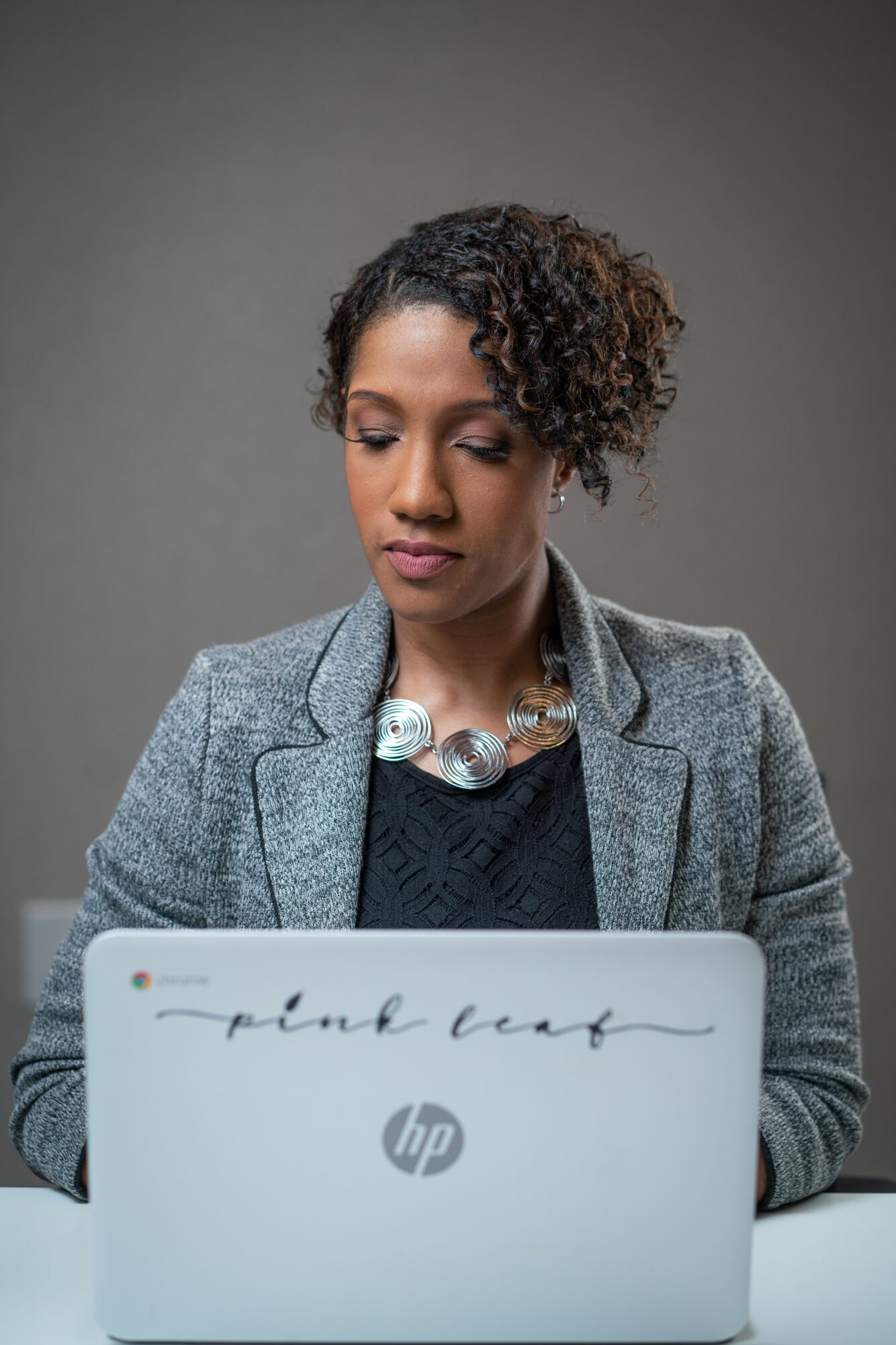 Woman with curly hair wearing a gray blazer and necklace, working on a laptop against a plain background.