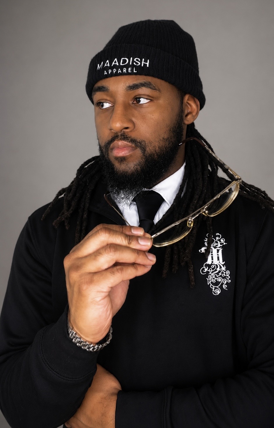 Man with a beard wearing a black beanie, black sweater, and holding glasses, looking to the side against a gray background.