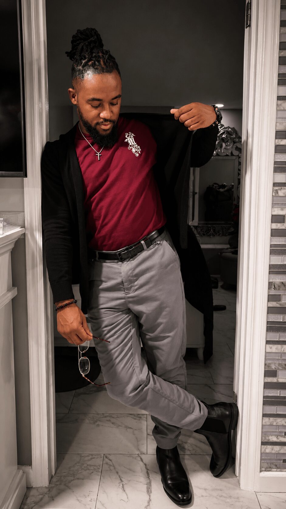 Man with a beard and braided hair, wearing a maroon shirt, gray pants, and black shoes, standing in a doorway.