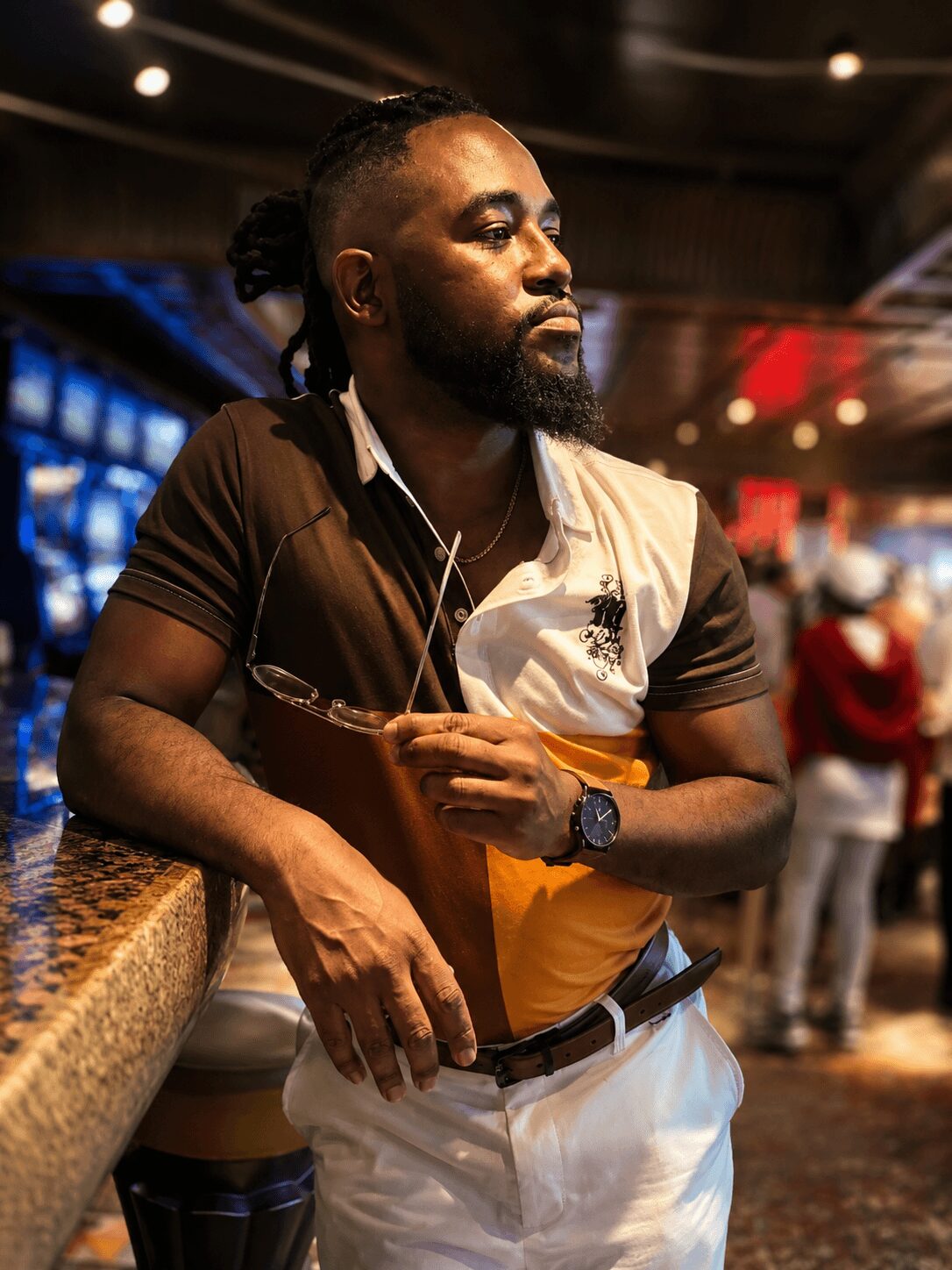 Man with beard and dreadlocks wearing a black and white shirt, holding a drink, standing in a lively indoor setting.