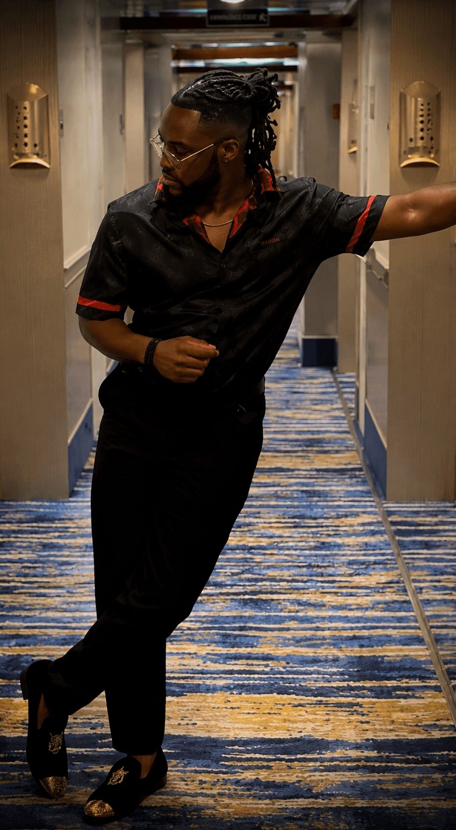 Person with braided hair and glasses leaning against a wall in a hallway, wearing a black outfit with red accents.