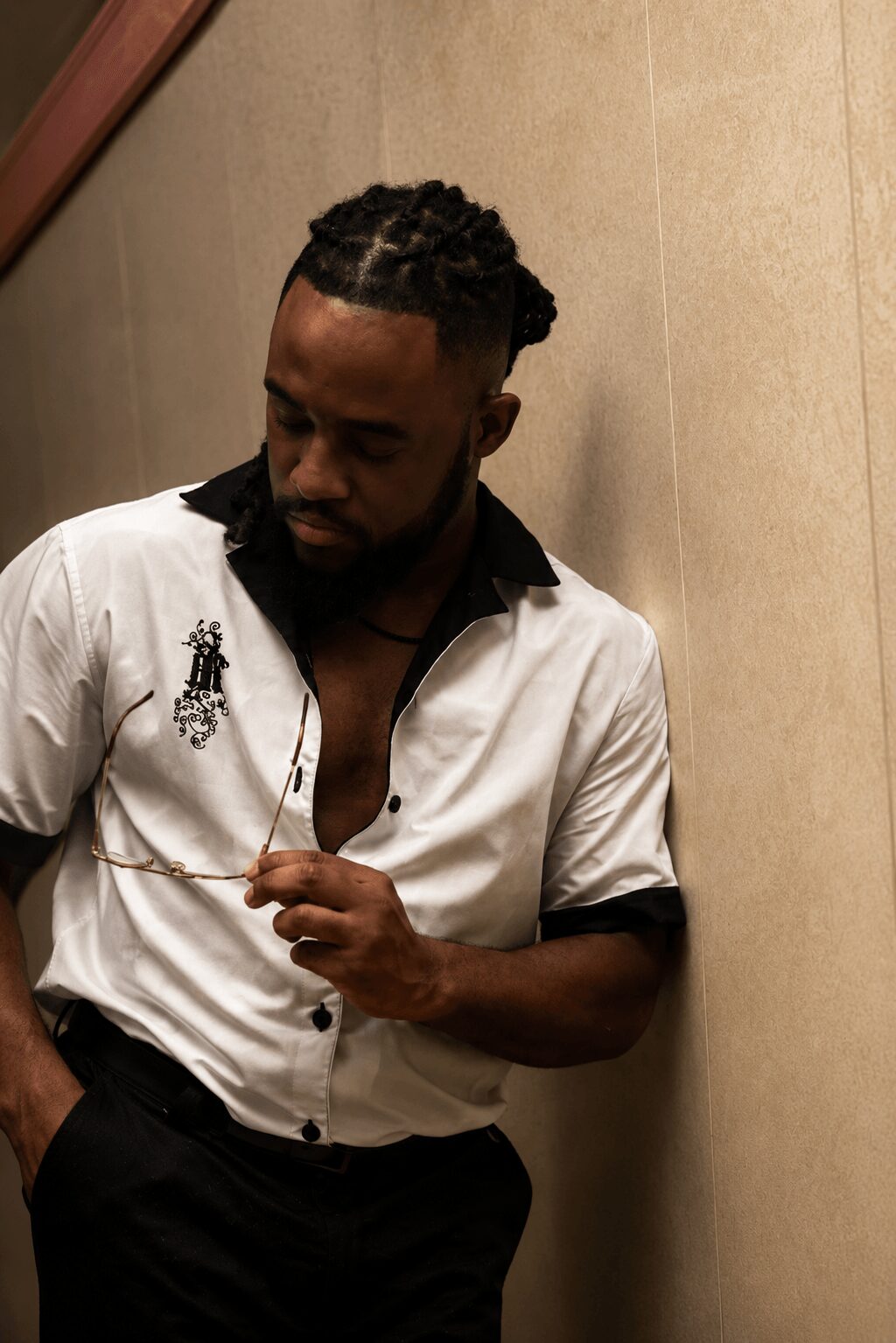 Man with styled hair and beard, wearing a white shirt with black collar, standing against a beige wall.