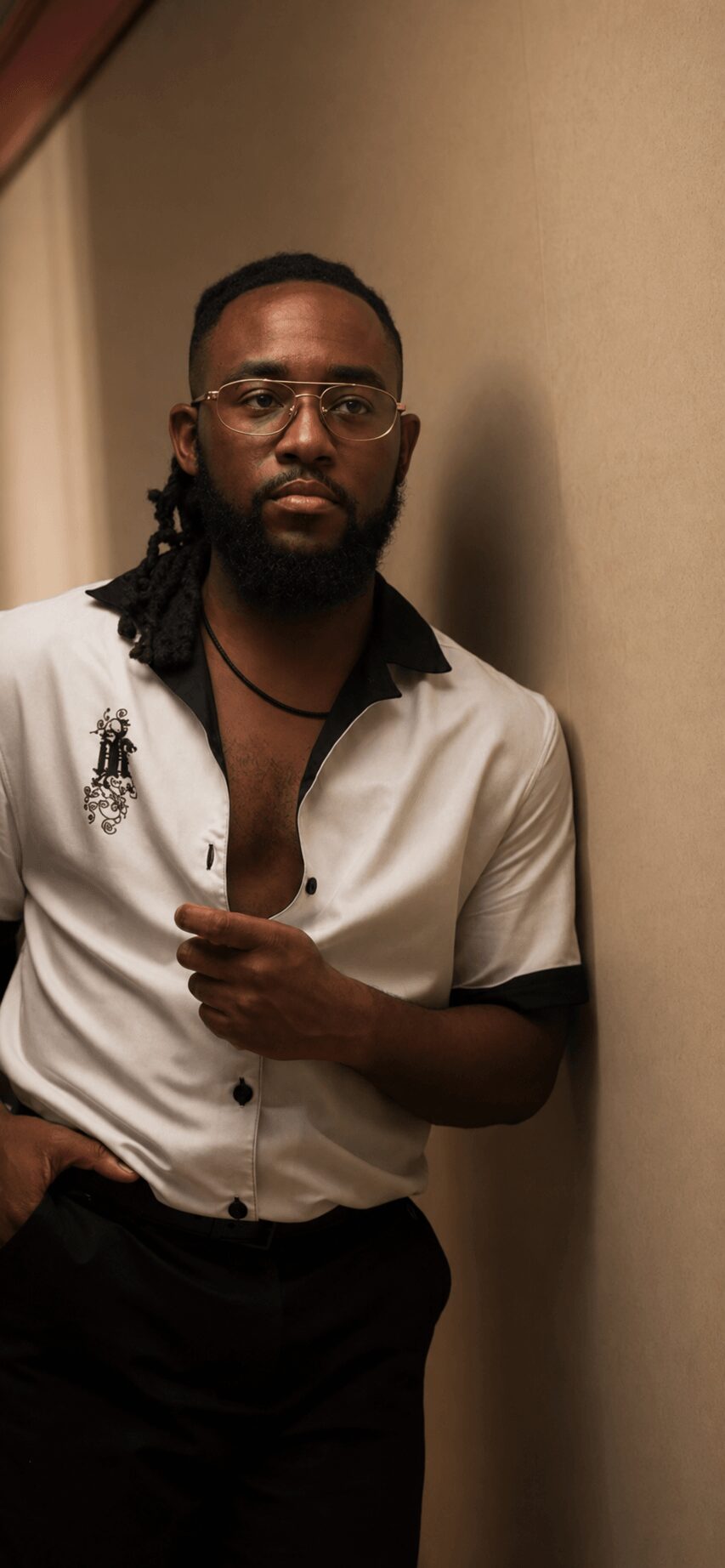 Man with glasses and dreadlocks wearing a white shirt, standing against a beige wall.