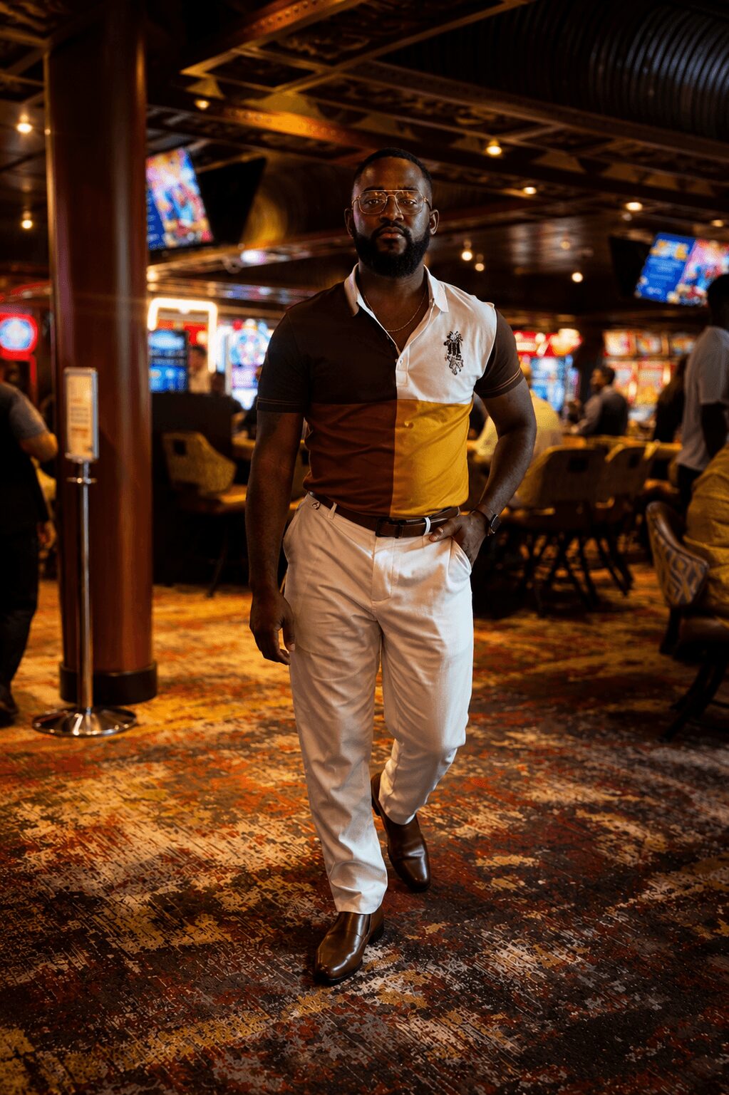 Man walking in a casino with slot machines and people in the background, wearing a multicolored shirt and white pants.