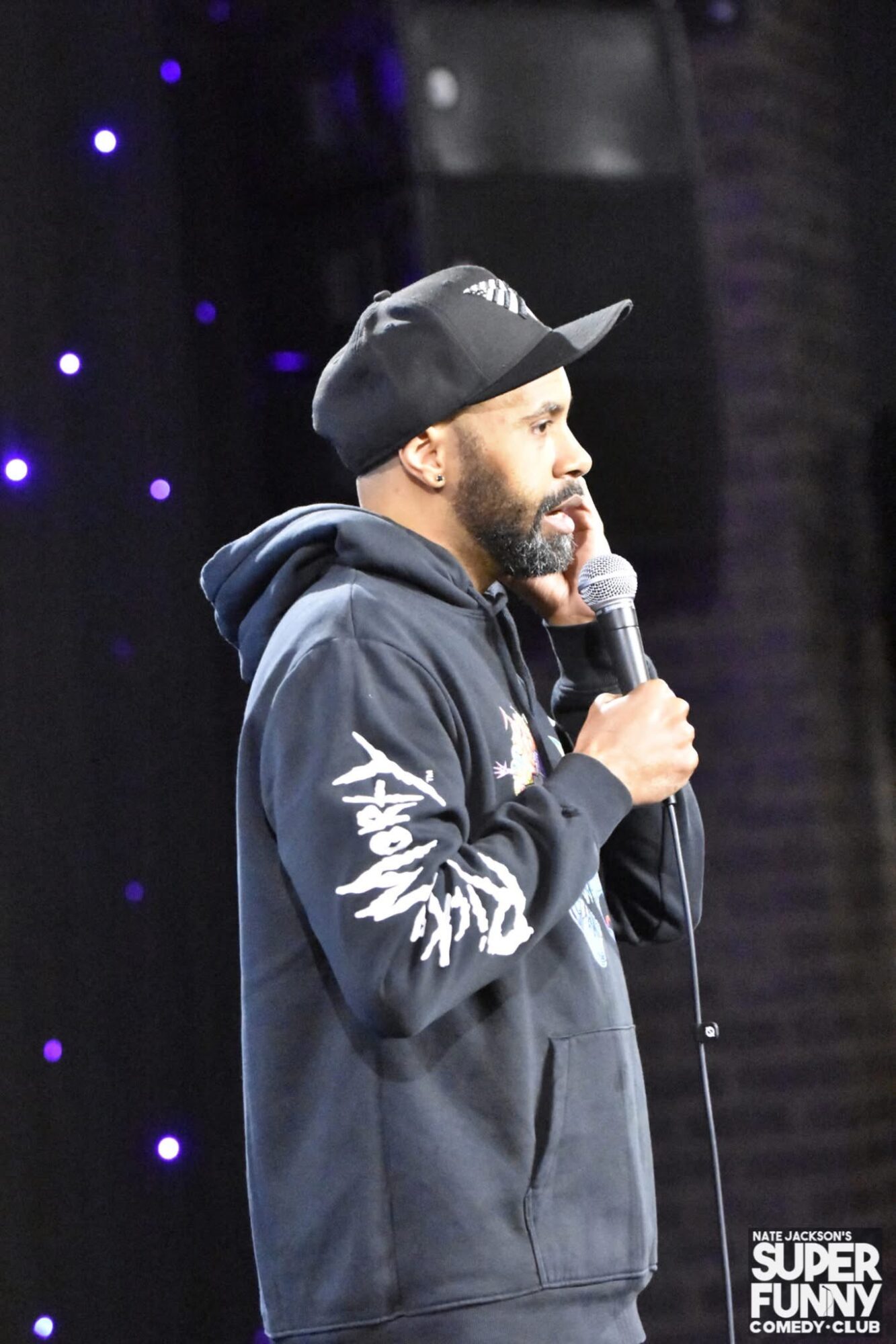 Man with beard wearing a black cap and hoodie, holding a microphone on stage with purple lights in background.