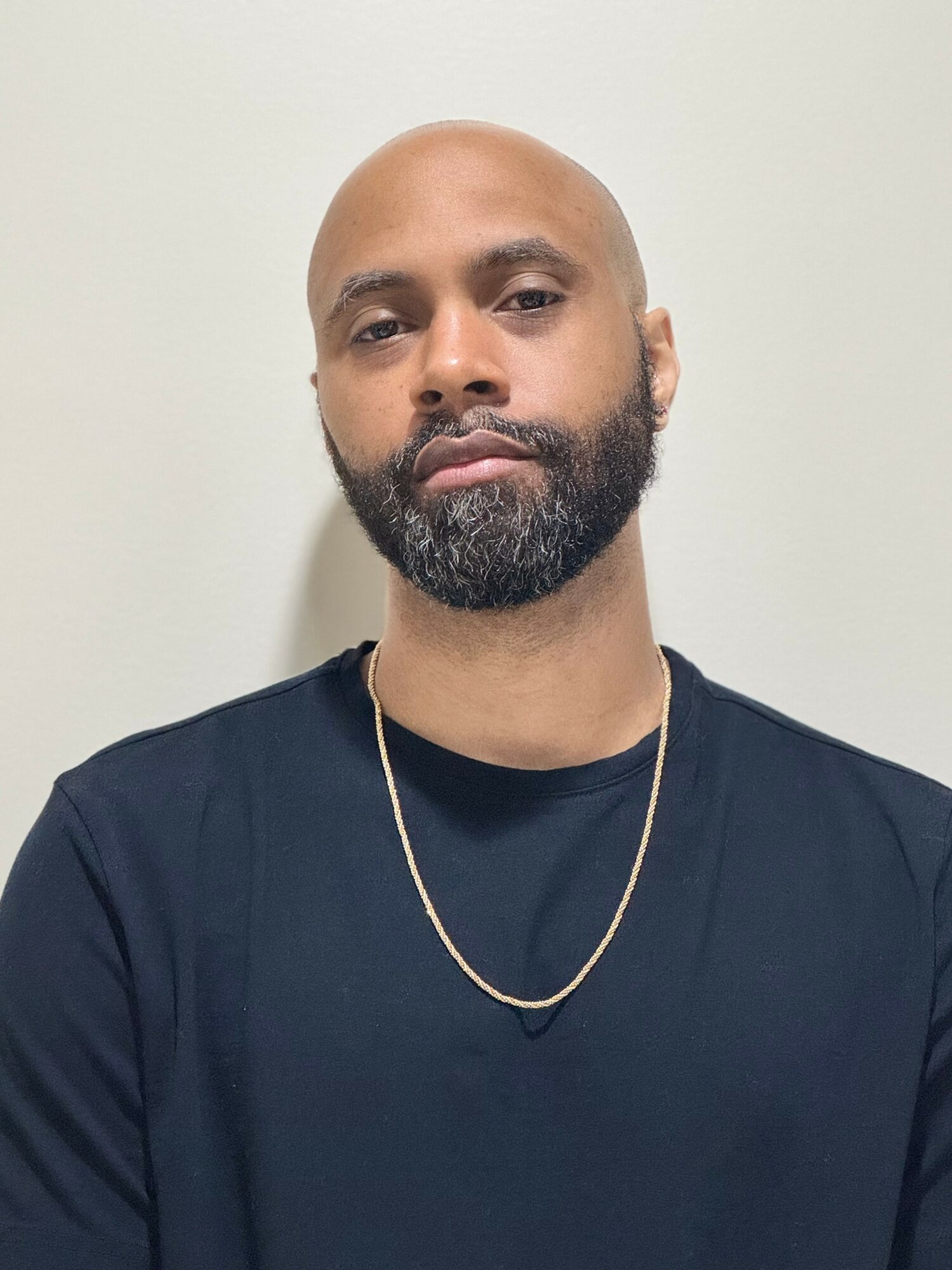 Bald man with beard wearing a black shirt and gold chain, looking at the camera against a plain background.