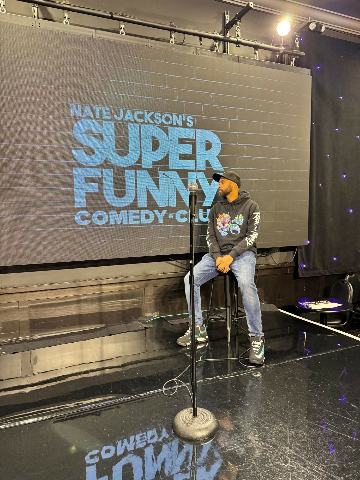 Person on stage with microphone, standing in front of a large screen displaying comedy show title, in a dimly lit venue.