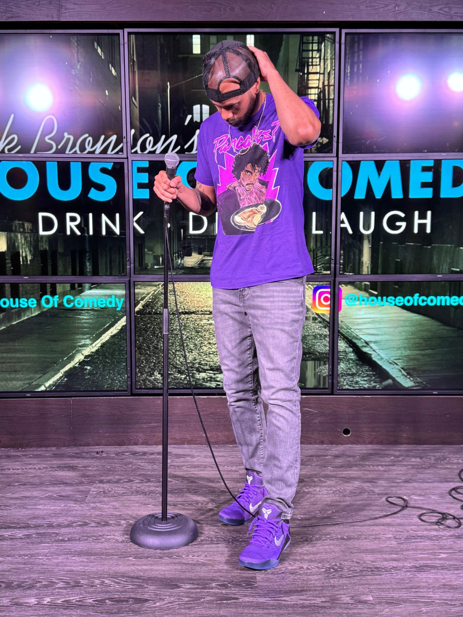 Person standing on stage with microphone, wearing purple shirt, gray pants, and purple shoes, holding a microphone stand.