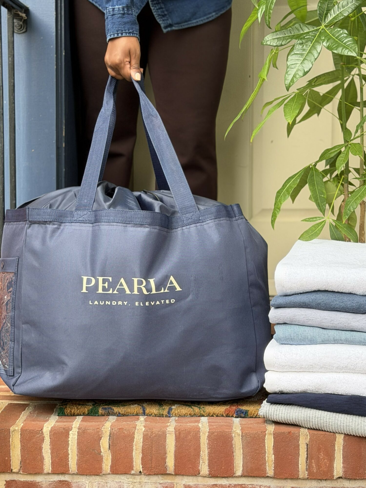 Person holding a large navy blue PEARLA laundry bag, standing next to a stack of folded towels and a green plant.