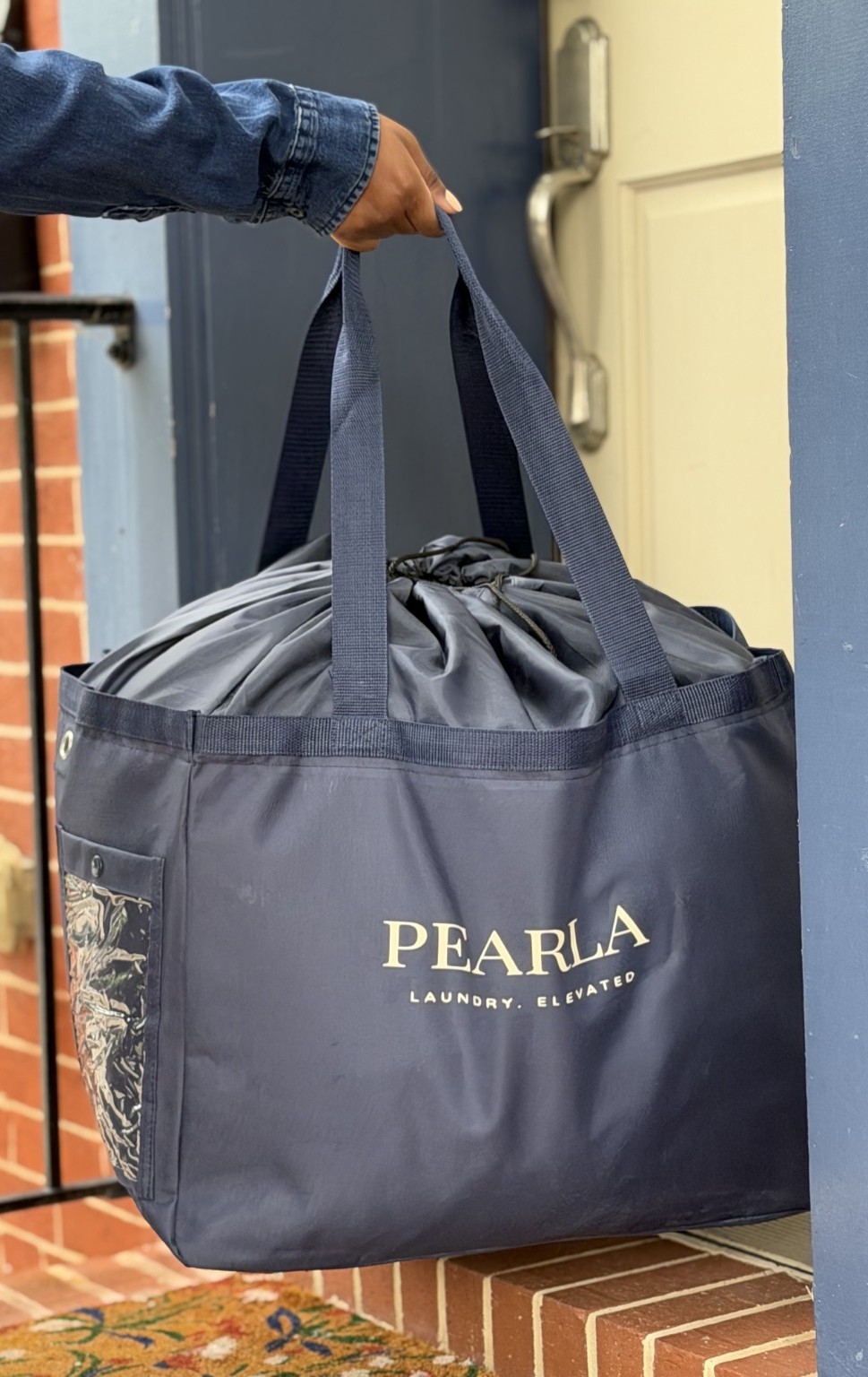 Person holding a large gray PEARLA laundry bag with handles outside a building, brick and door visible in background.