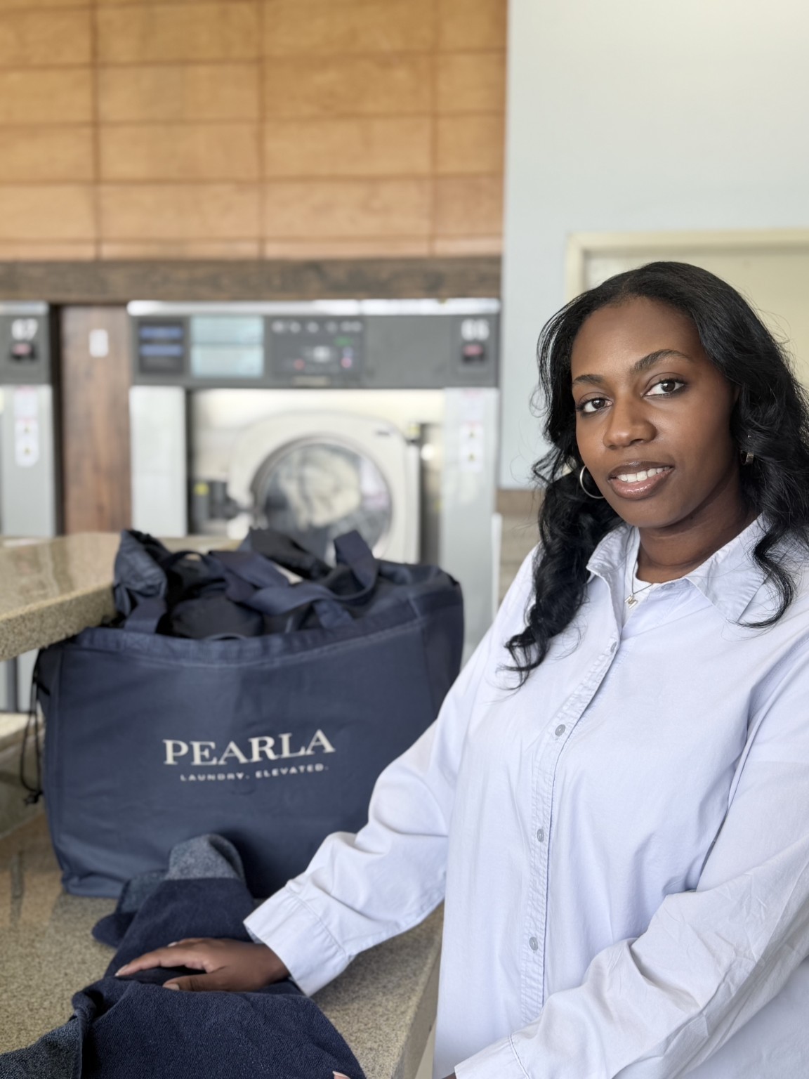 Woman with long dark hair in a white shirt sitting at a counter, laundry bag labeled PEARLA nearby, laundry machines in background.