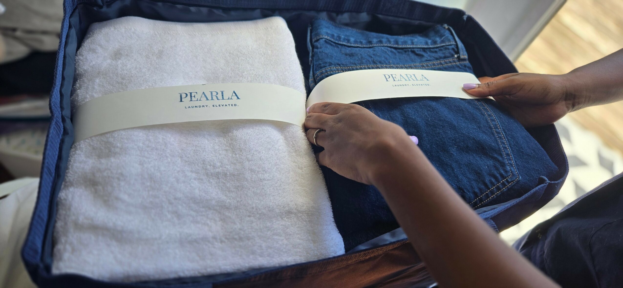 Person packing white towel and blue jeans into a suitcase, hands visible, sunlight in background.
