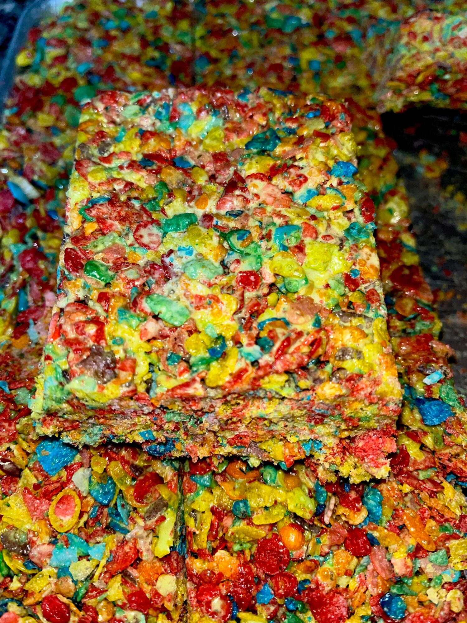 Colorful cereal treat with various small, brightly colored cereal pieces, cut into a square, with some crumbs around it.