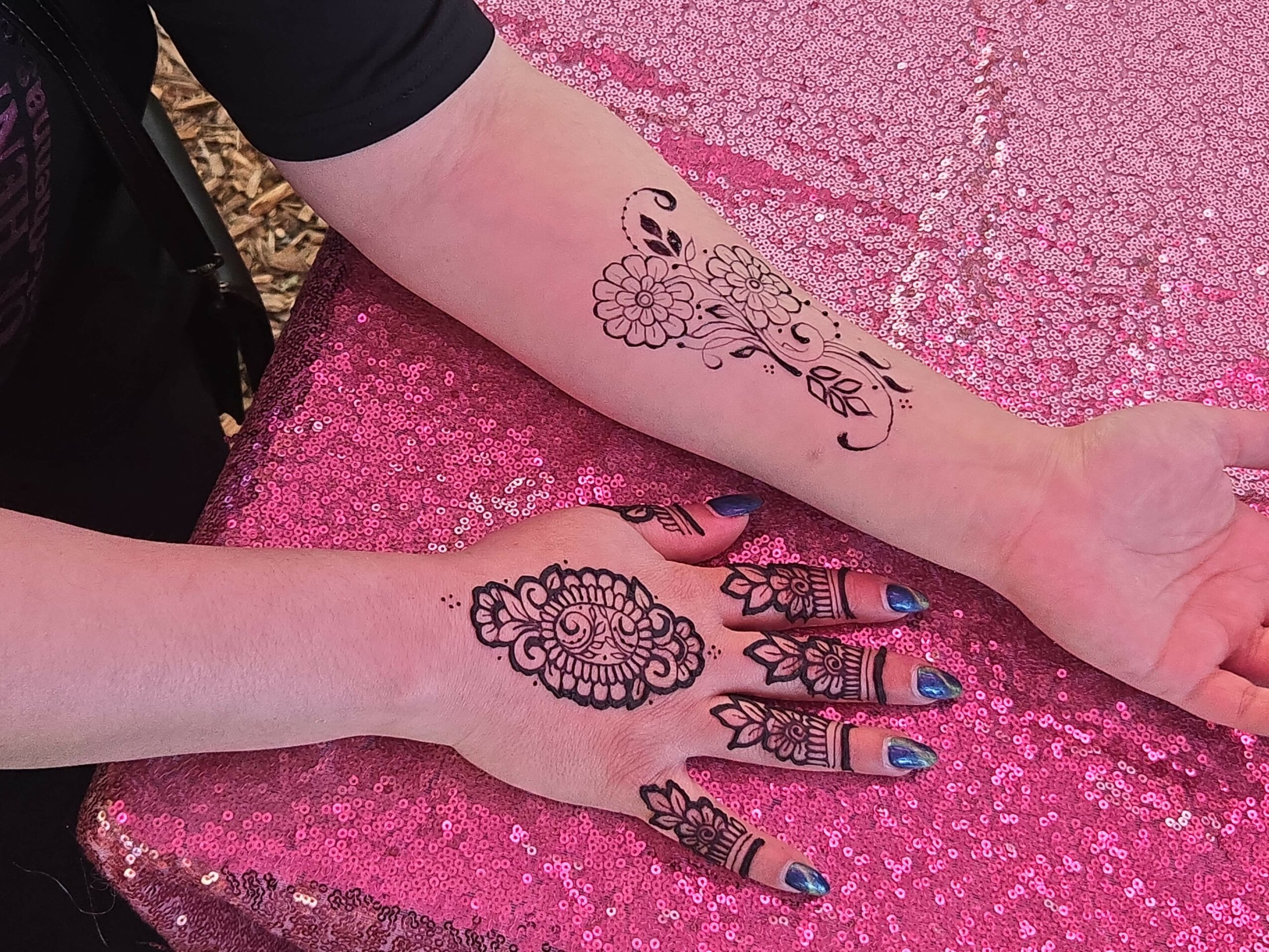 Two arms with tattoos resting on a pink, glittery surface. One arm has a floral tattoo, the other a mandala design.