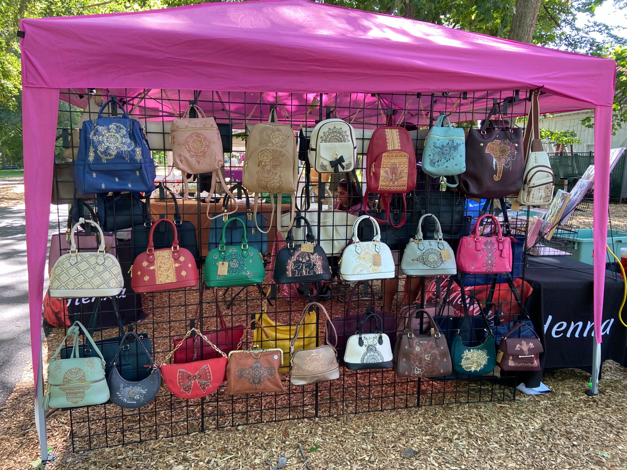 Display of various handbags on a wire grid under a pink canopy outdoors, with trees and grass in the background.