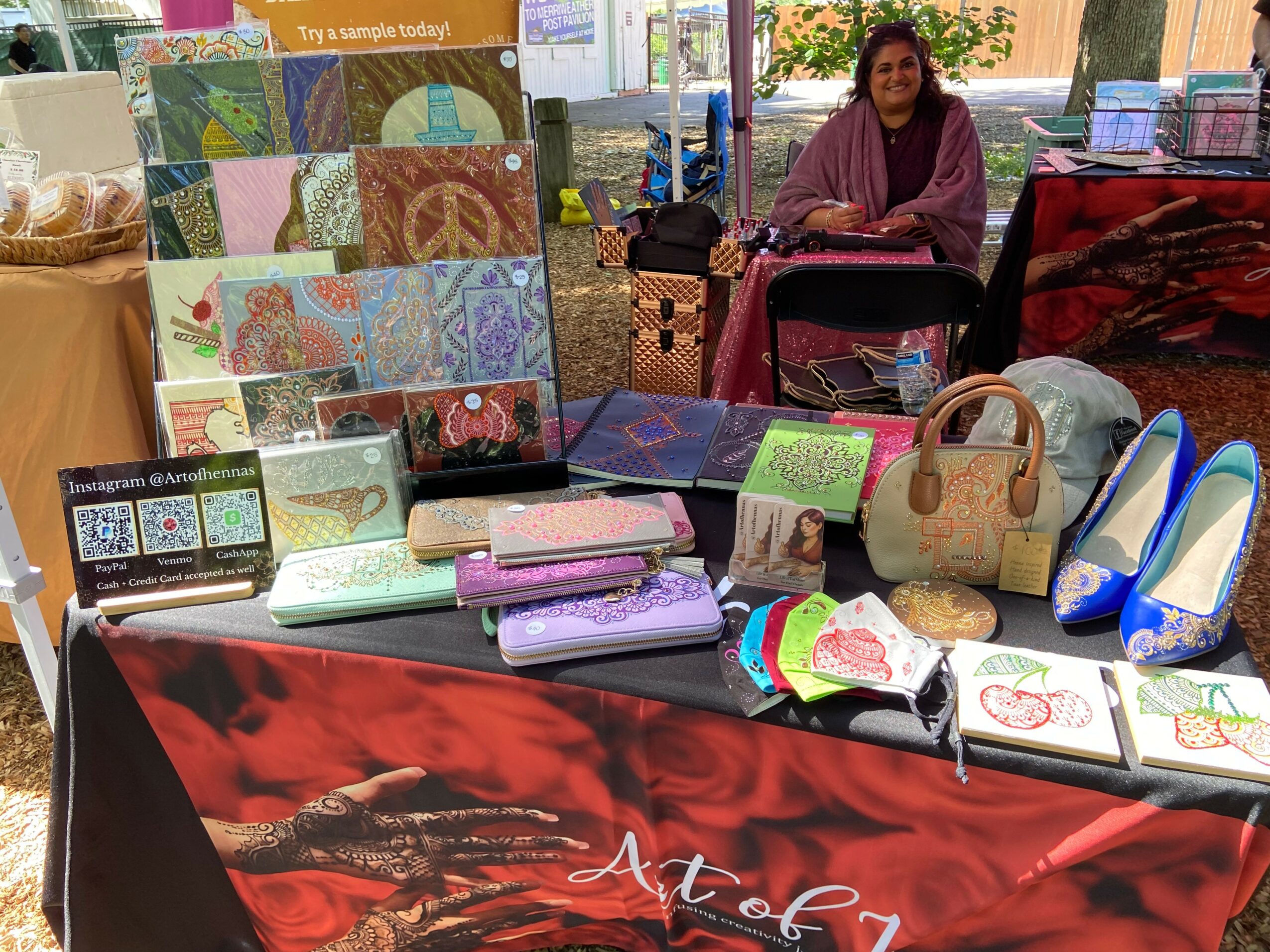 Woman sitting behind a table displaying colorful handmade fabric items, bags, and artwork at an indoor market.
