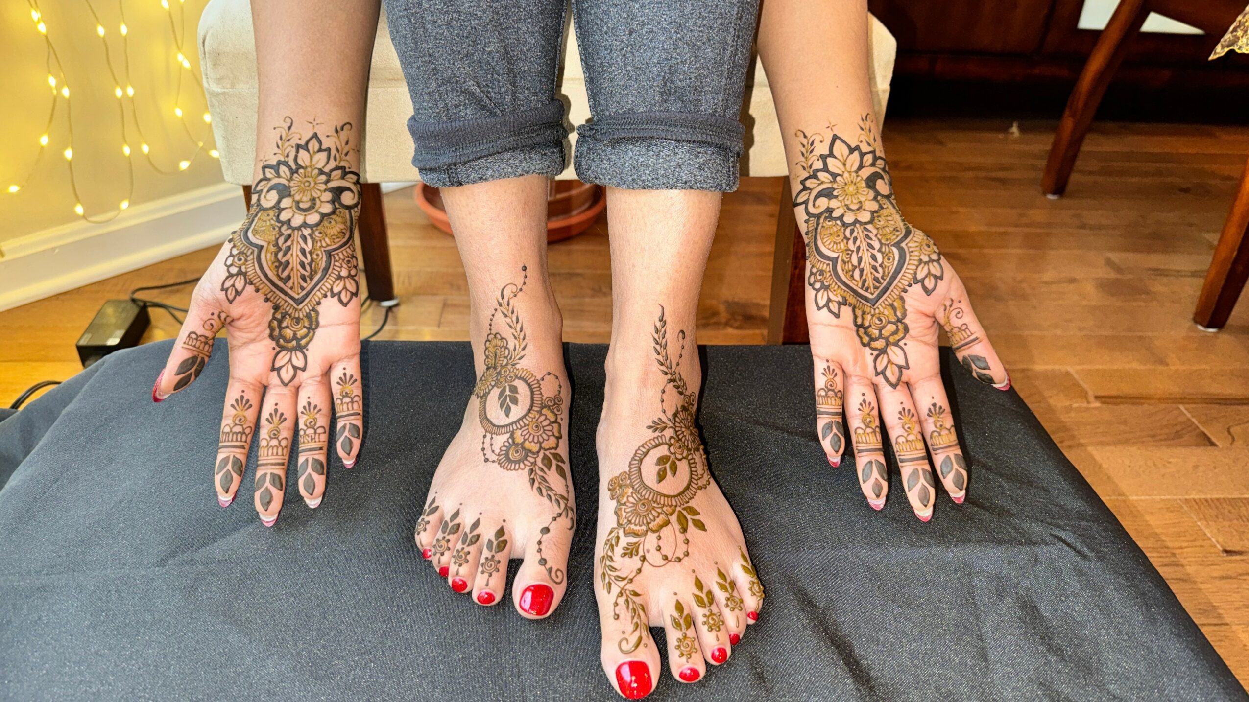 Person with henna designs on hands and feet, red nail polish, standing on a black surface, wooden floor in background.
