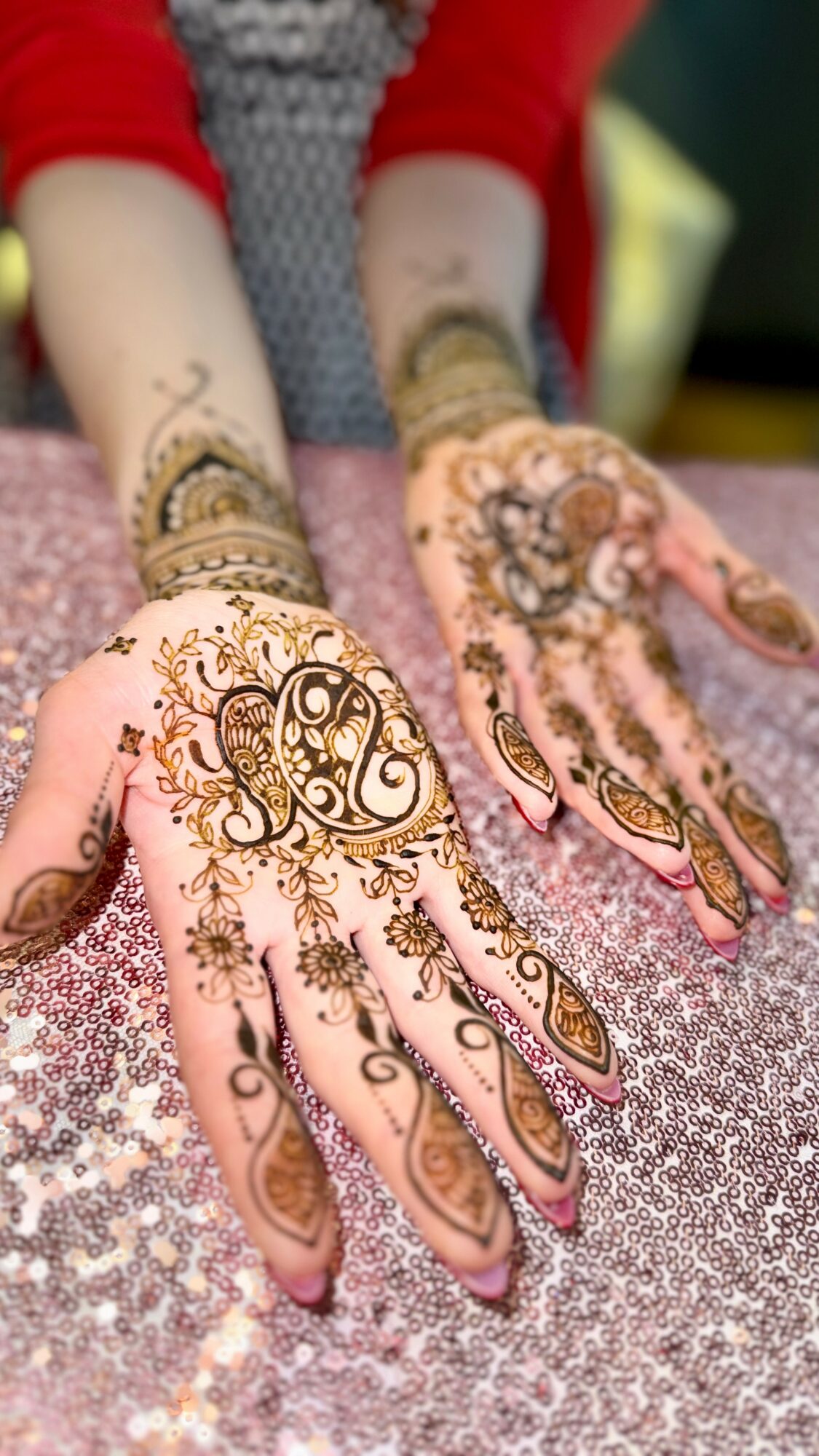 Two hands with intricate henna designs resting on a sparkly surface, with a person wearing a red sleeve in the background.