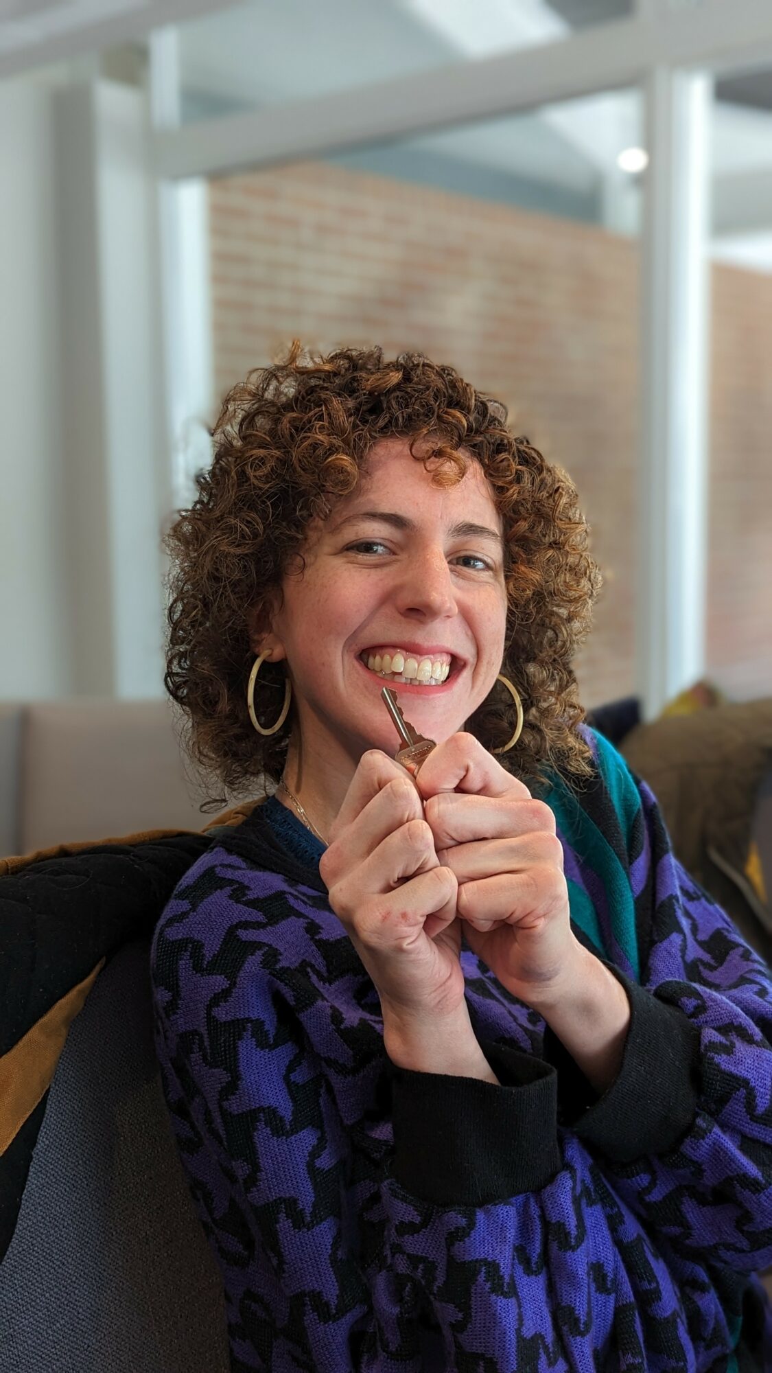 Woman with curly hair smiling, holding a small object, wearing a purple patterned jacket and hoop earrings, indoors.