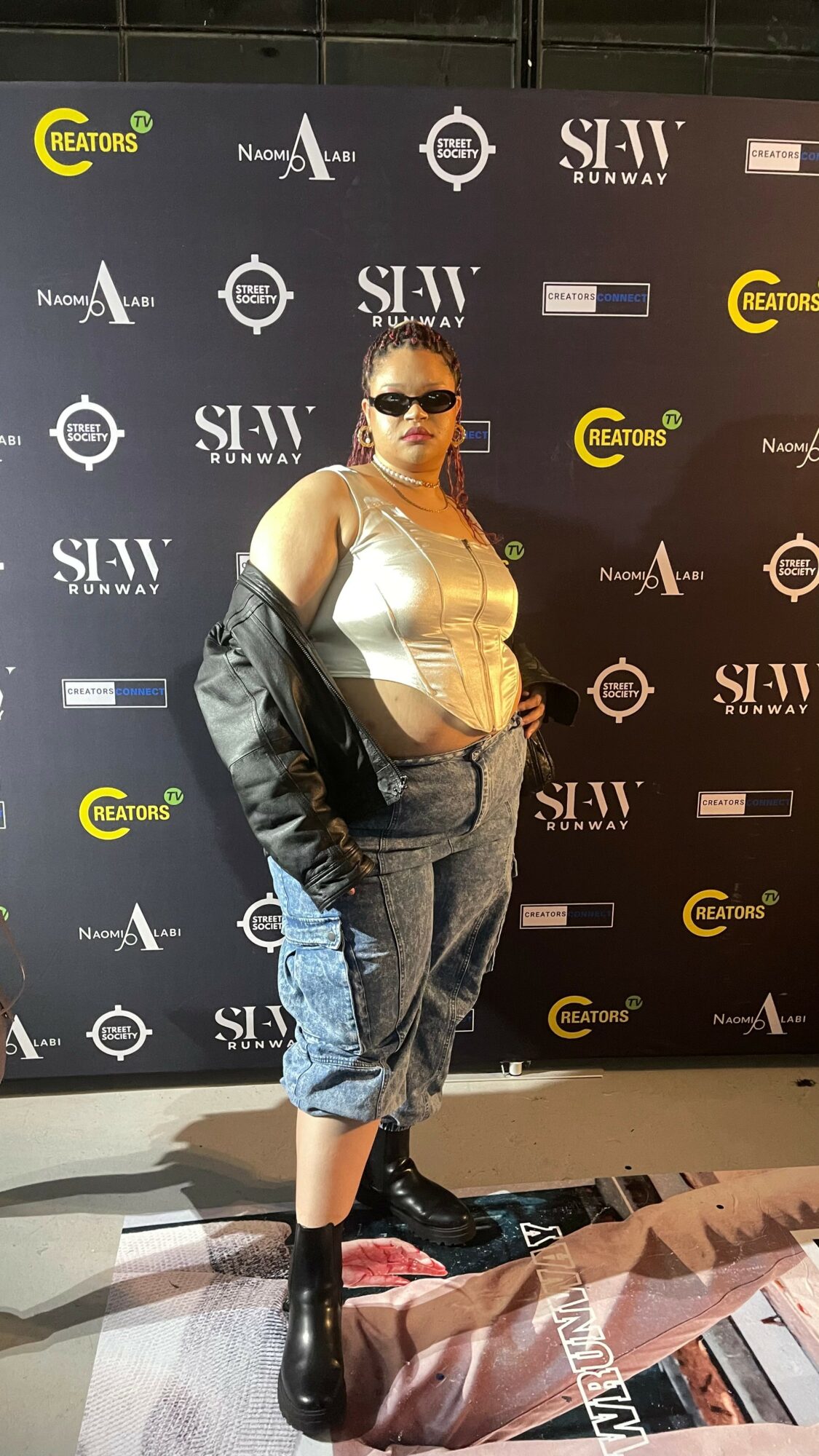 Person with sunglasses and a black jacket over a beige top, standing in front of a black backdrop with logos.