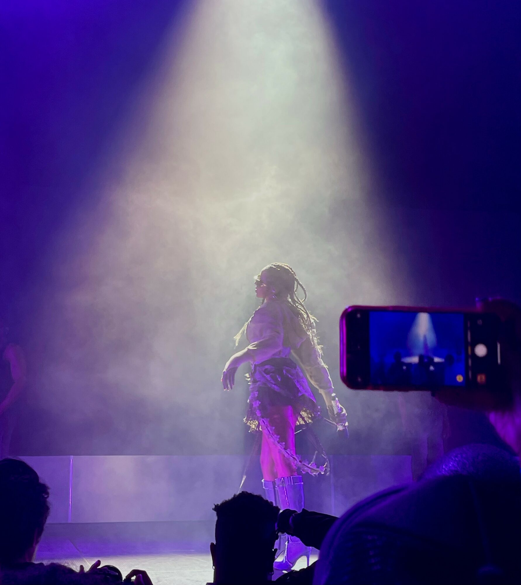 Person on stage under spotlight, facing away, with audience and a person taking a photo in foreground.
