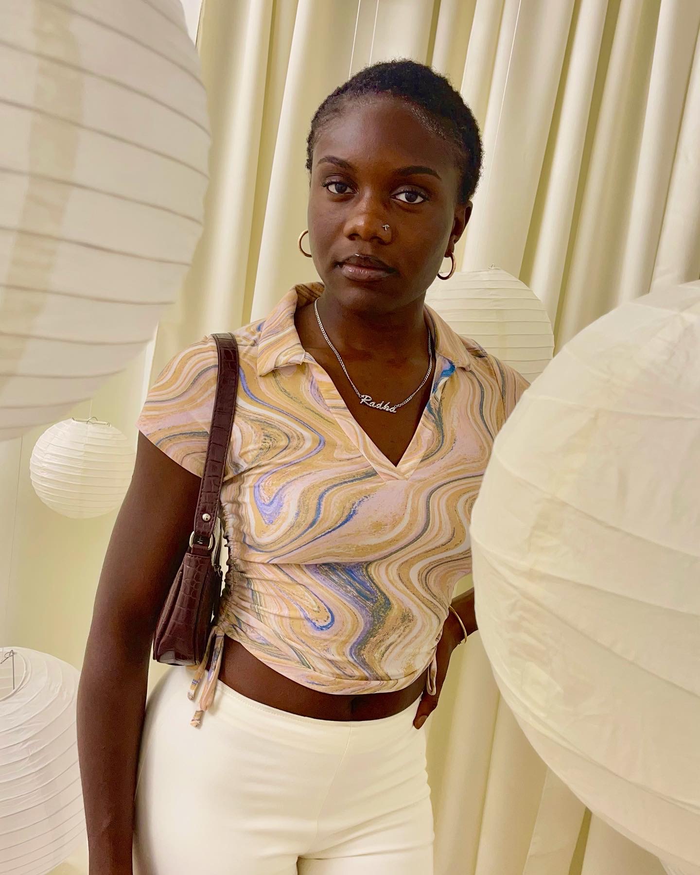 Young woman with short hair wearing a colorful patterned crop top and white pants, standing indoors with cream curtains in background.