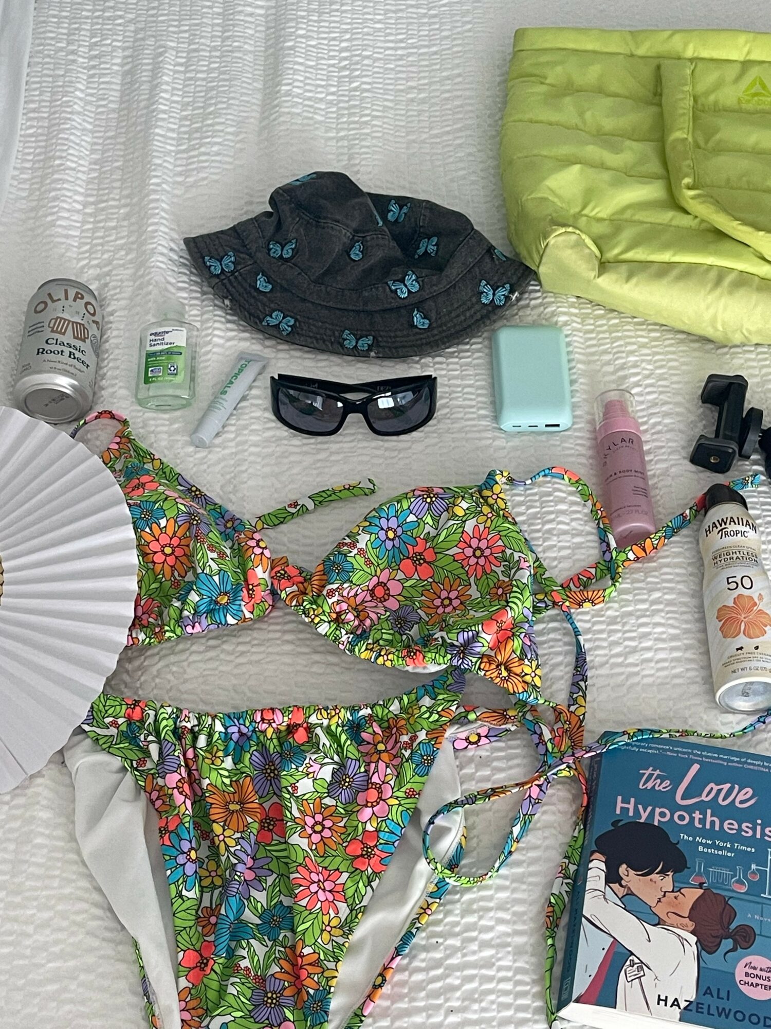 Beach items including a floral bikini, sunglasses, sunscreen, a fan, a hat, and a book on a white surface.
