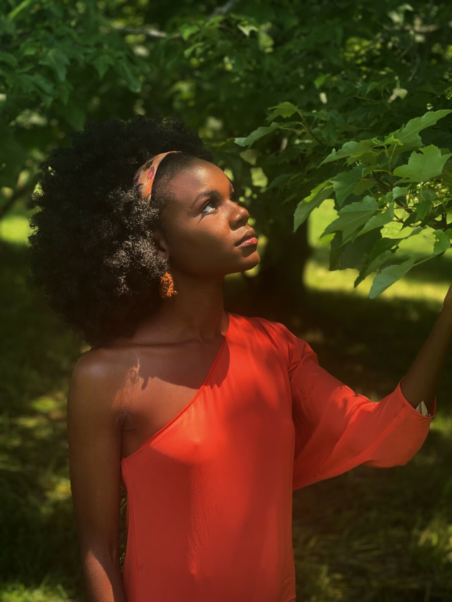 Young woman with curly hair wearing a red dress, standing outdoors among green leaves, looking upward at the foliage.