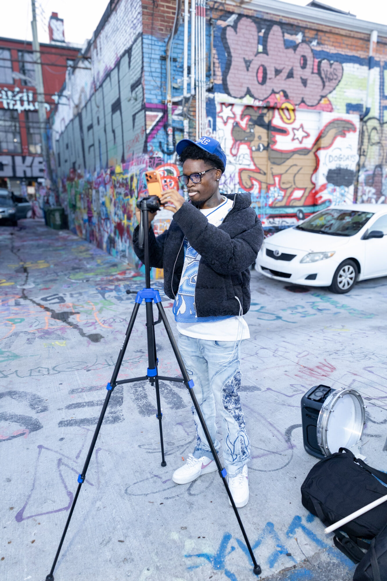 Young man with glasses and a cap taking a photo with a camera on a tripod in an urban alley with colorful graffiti walls, a white car, and street art.