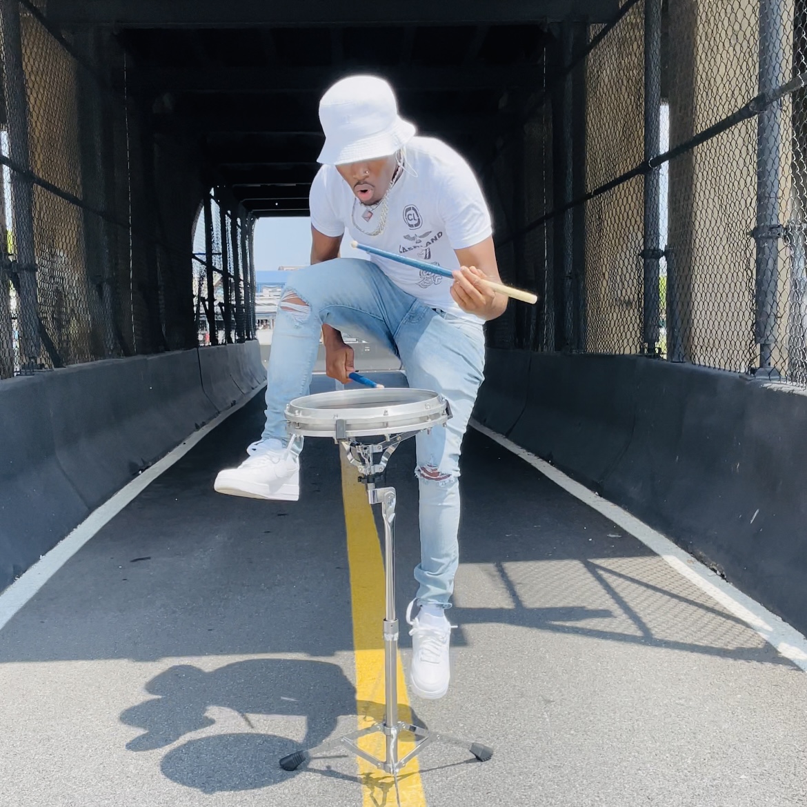 Person playing drum on street, wearing white hat and shirt, light blue jeans, and white shoes, under a bridge.