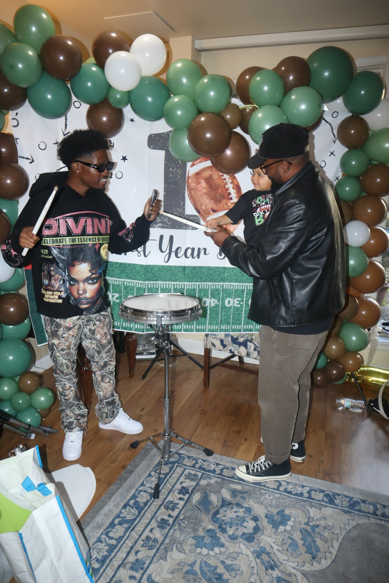 Person speaking into microphone at celebration with green, white, and brown balloons, and a woman holding a photo, in a decorated room.