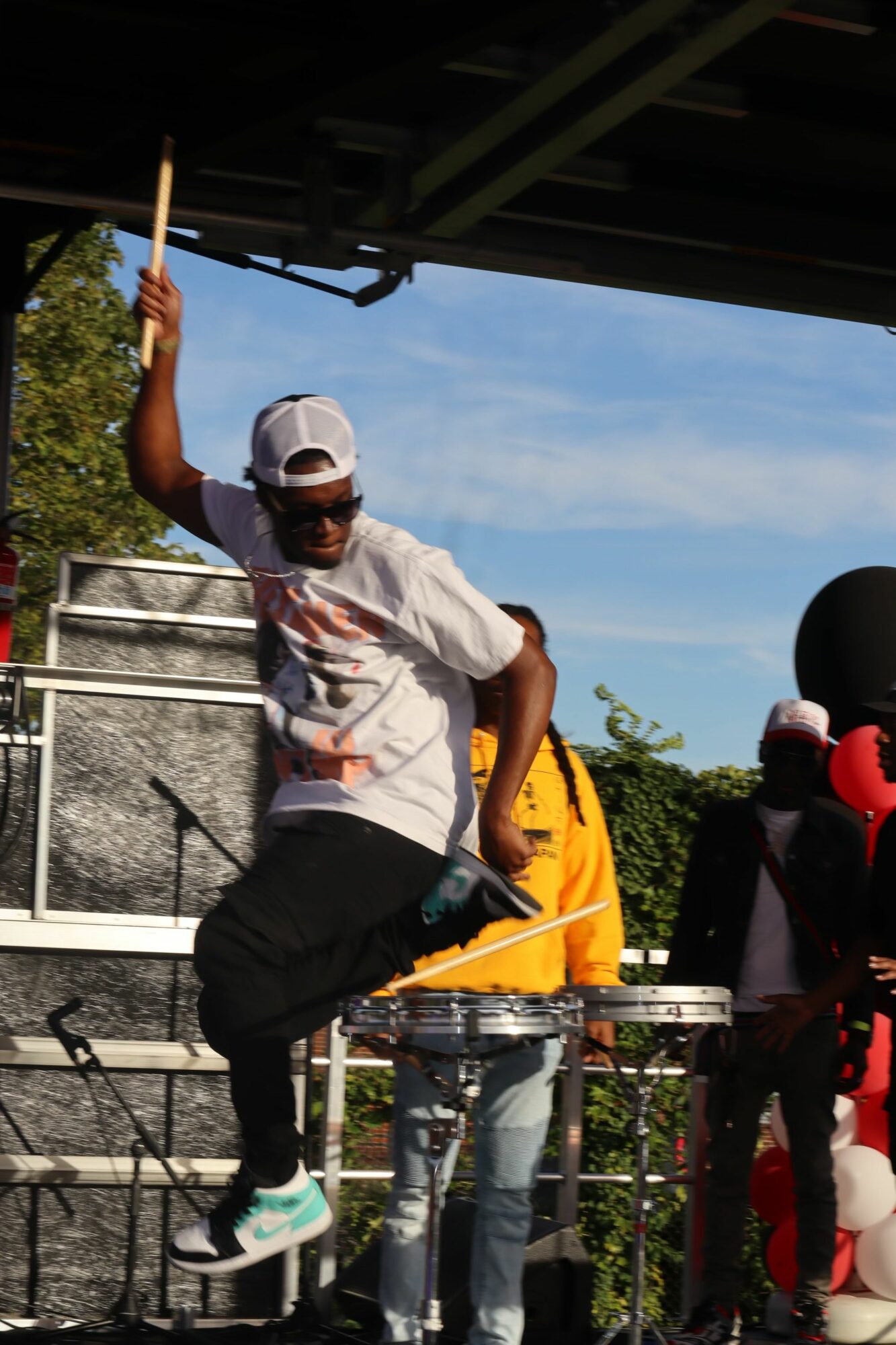 Person on stage holding a drumstick, wearing a cap and white shirt, with a blue sky background.