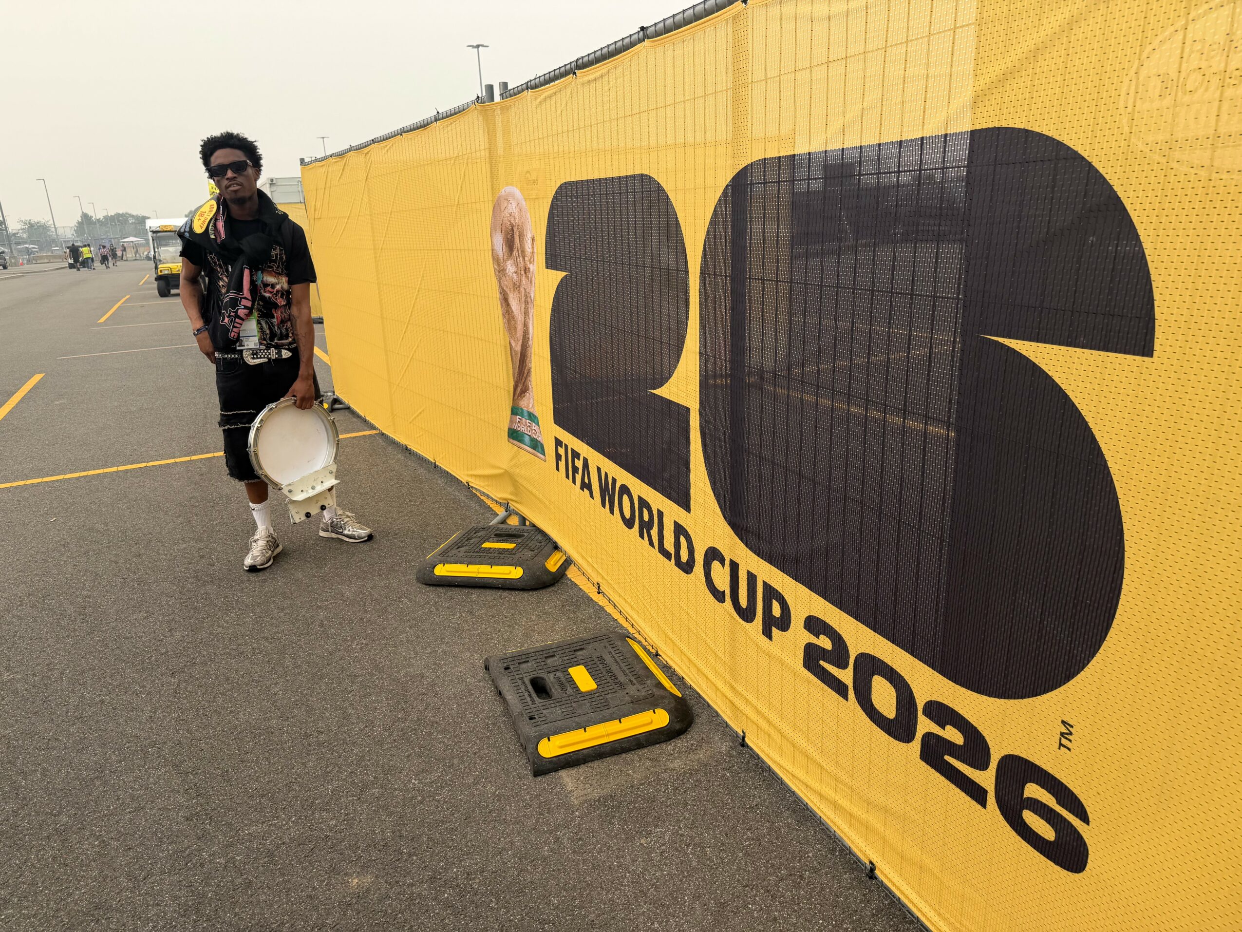 Person standing next to a yellow FIFA World Cup 2022 banner on a street, holding a white object, with barriers nearby.