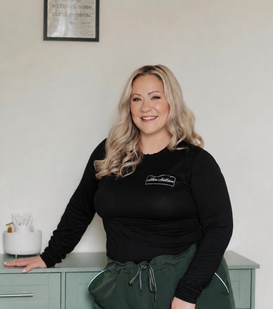 Smiling woman with blonde hair in a black top standing next to a light green cabinet, with a white wall and framed picture behind her.
