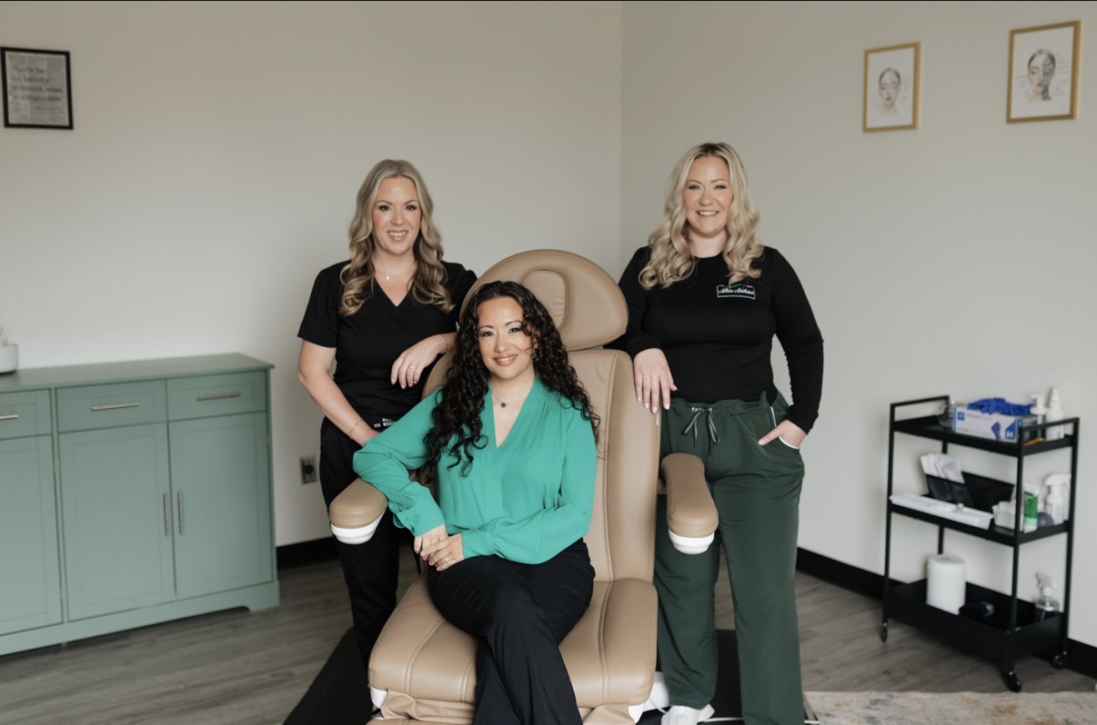 Three women in a room with light-colored walls, two standing and one sitting on a chair, smiling.