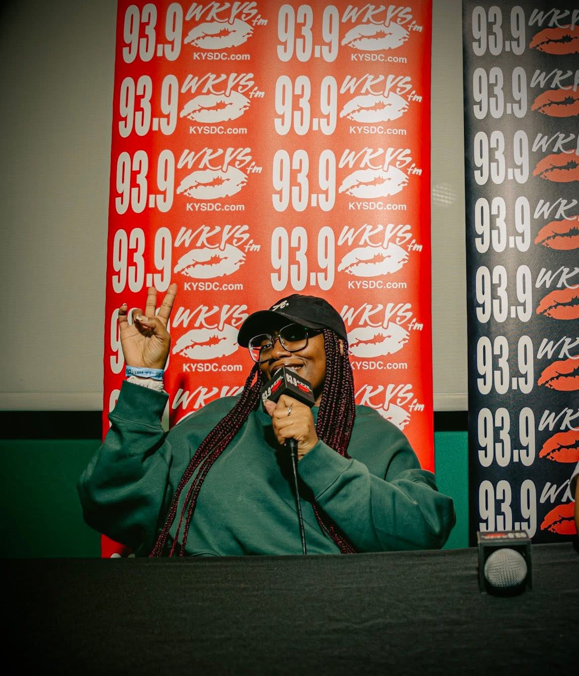 Person with long braided hair, glasses, and a cap, speaking into a microphone at a radio station, background with red and black logos.