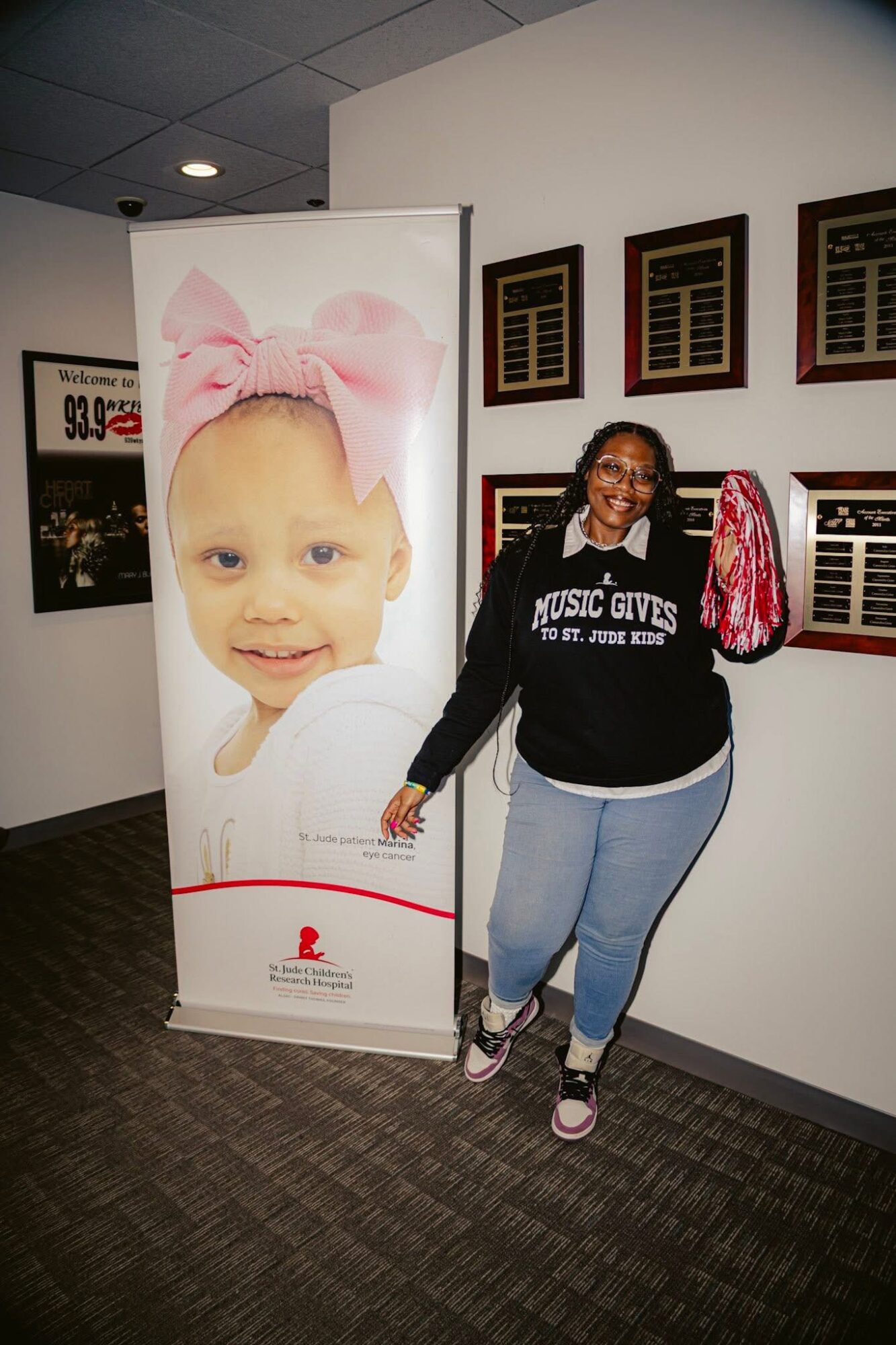 Person standing next to a large poster of a smiling child with a pink bow on her head, inside a building with framed items on the wall.